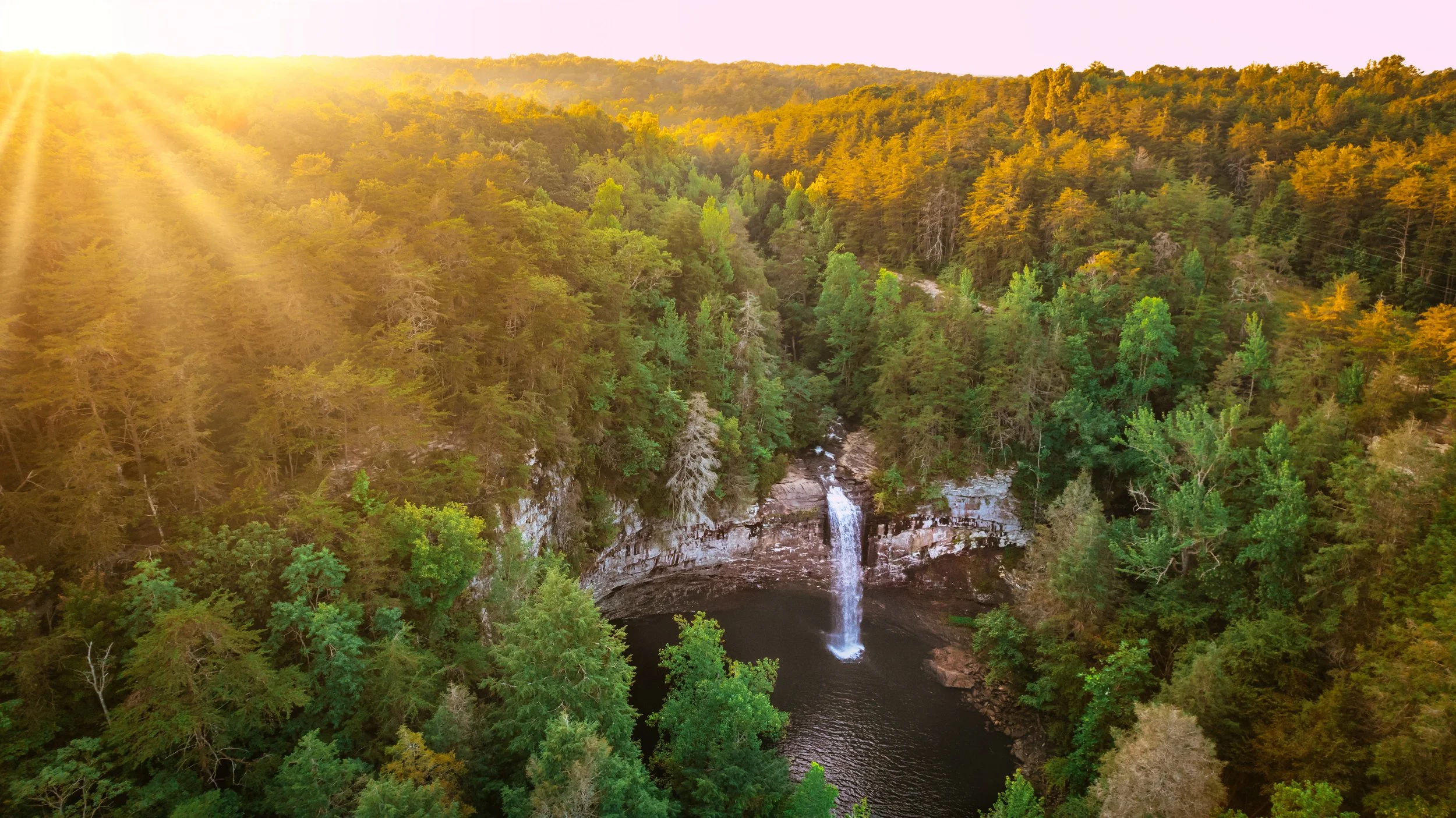 Aerial view of a waterfall in a lush forest with autumn colors, bathed in warm sunlight.