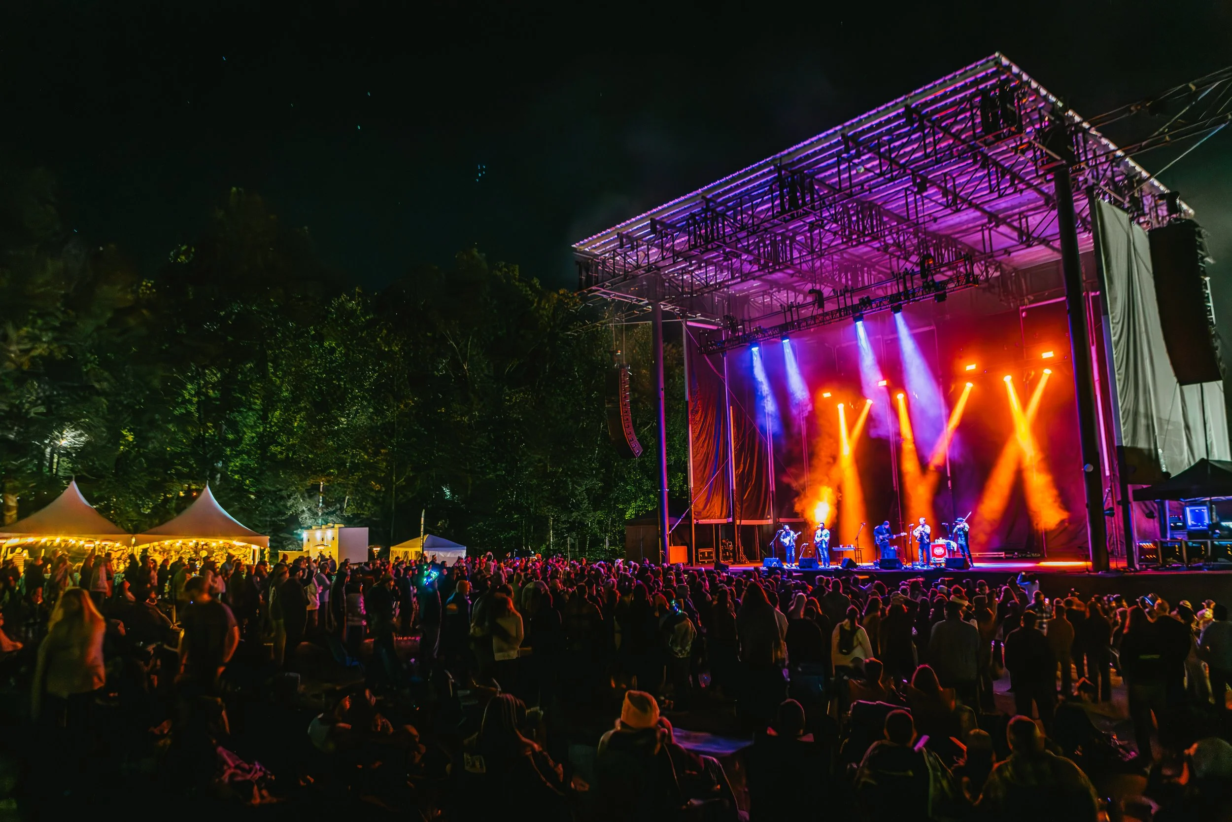 Nighttime outdoor concert with a large crowd watching a band perform on stage with vibrant orange, purple, and blue lighting; trees and tents are visible in the background.