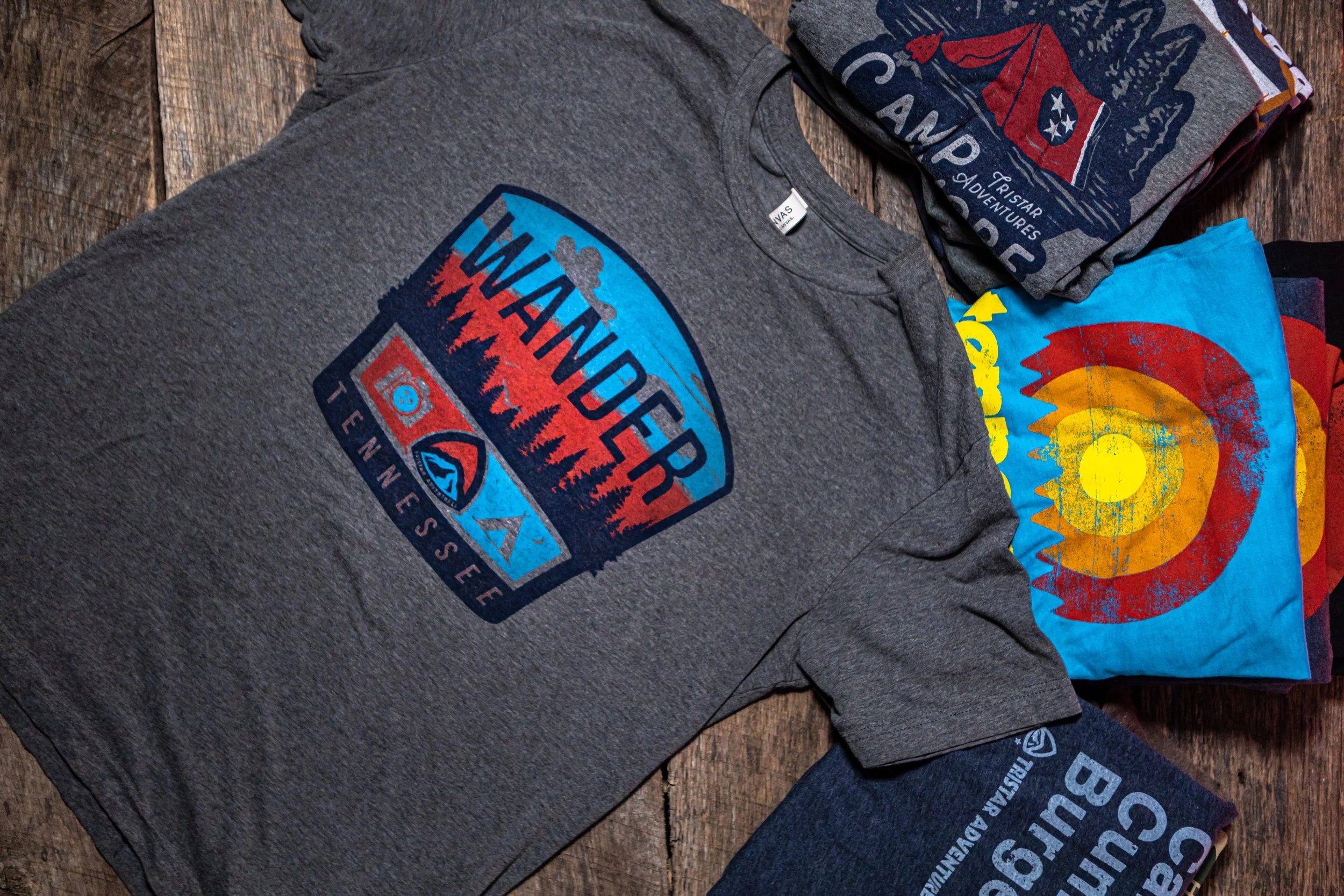 A display of folded T-shirts on a wooden surface, featuring designs related to outdoor and adventure themes, including a mountain scene with the text "Wander Team" and colorful, abstract patterns.