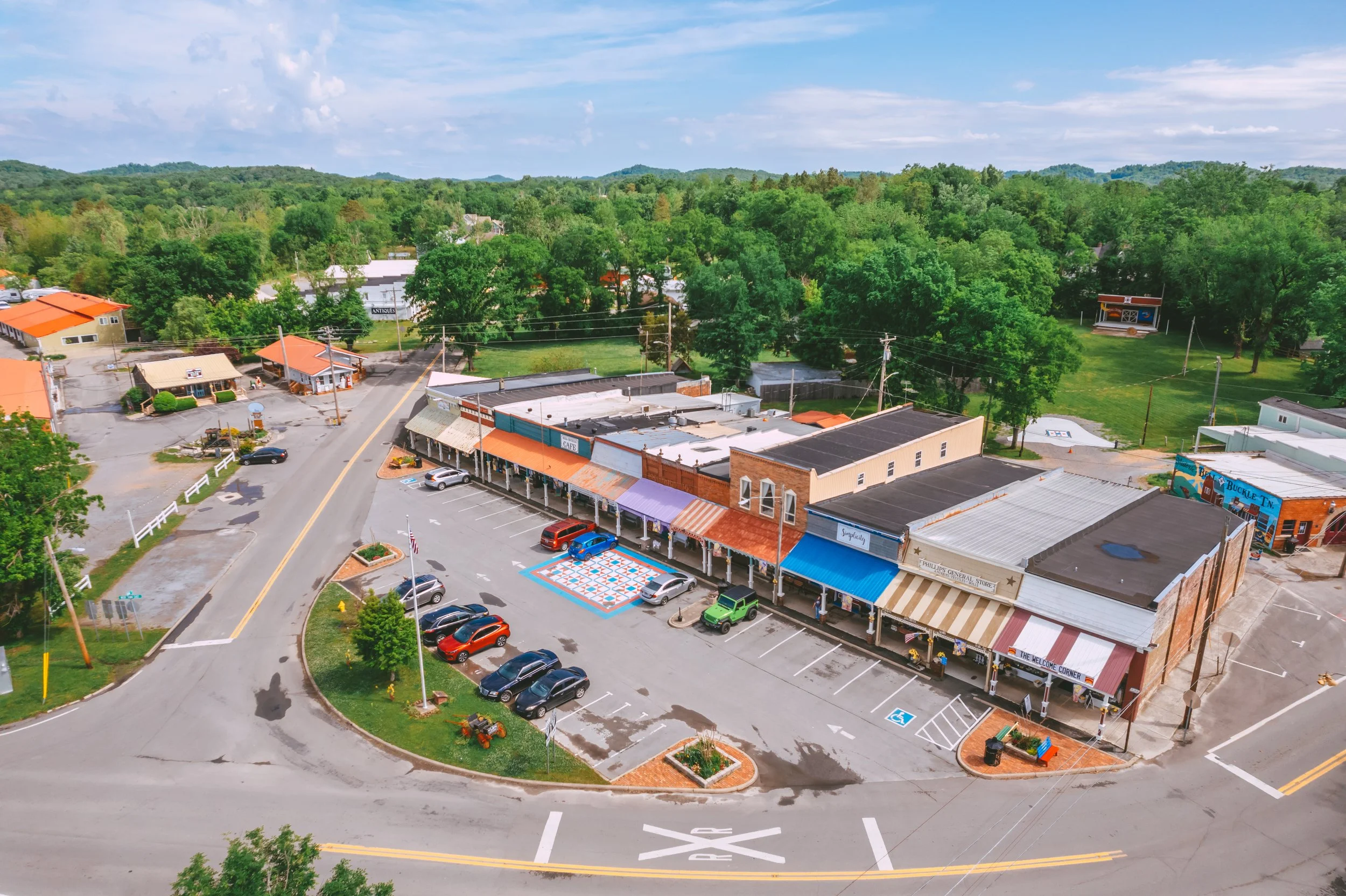 An aerial view of a small town shopping area with shops, parked cars, and green trees in the background under a partly cloudy sky.