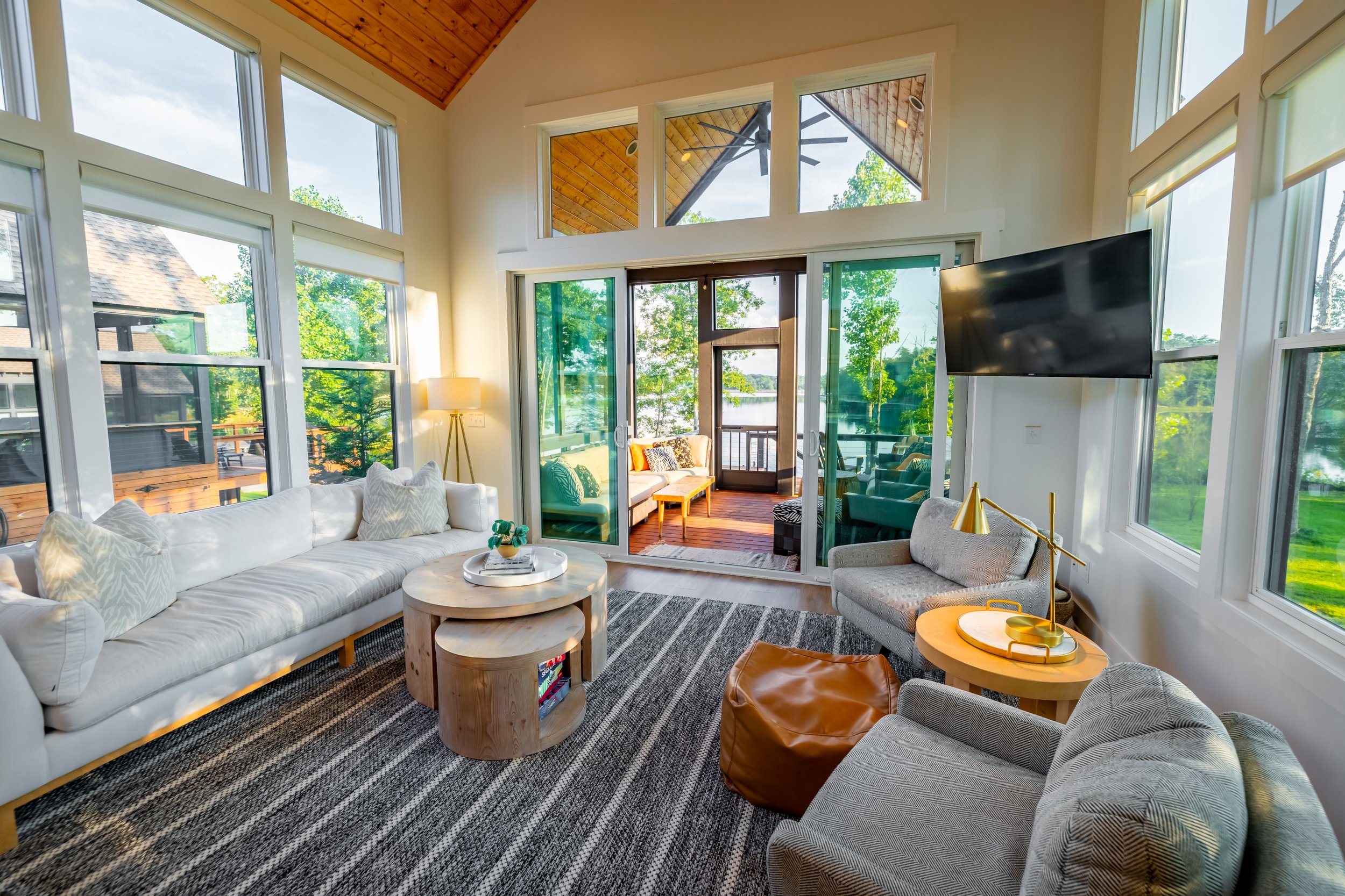Bright living room with large windows and sliding glass doors leading to a balcony with outdoor seating, featuring a white sofa, gray armchair, wooden coffee tables, a wall-mounted TV, and a patterned rug.