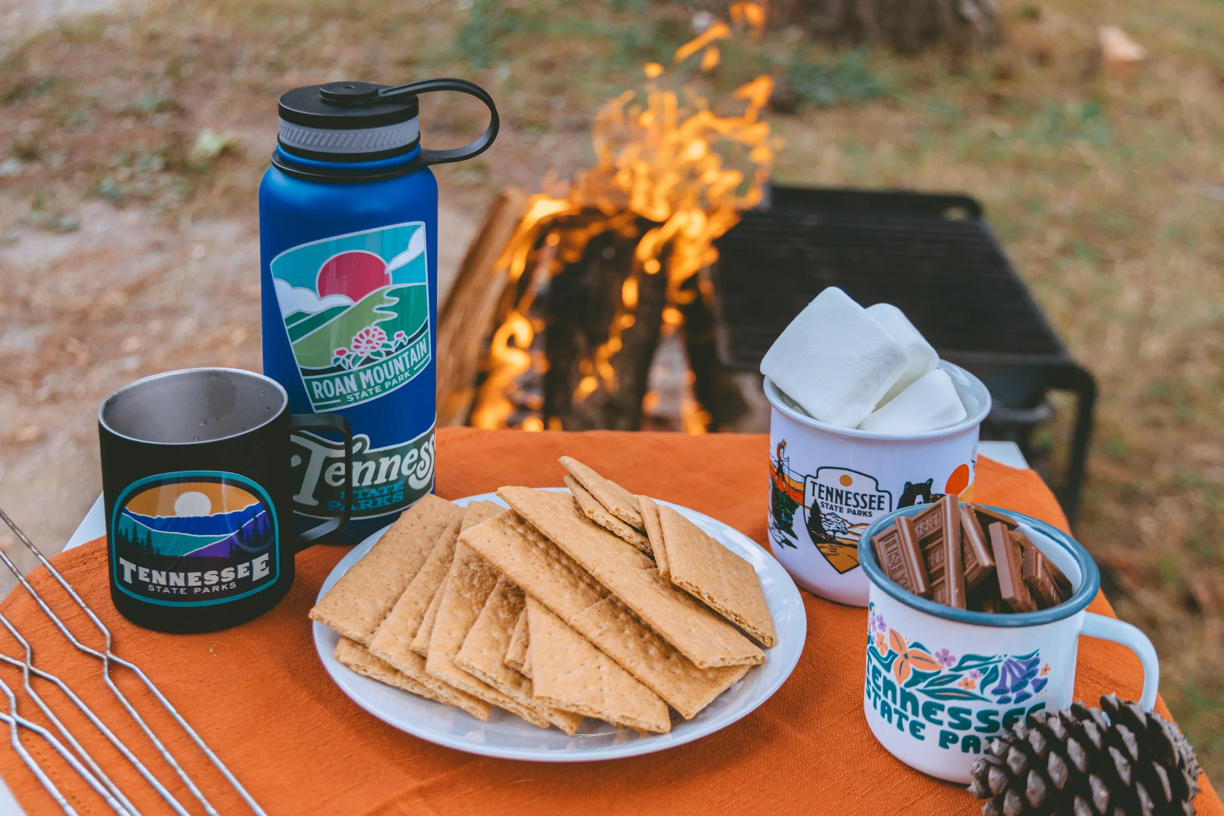 A camping scene with a table set with wheat crackers, marshmallows in a Tennessee State Parks cup, chocolate in a Tennessee State Parks mug, a blue Tennessee State Parks water bottle, and a black Tennessee State Parks mug, all in front of a lit campf