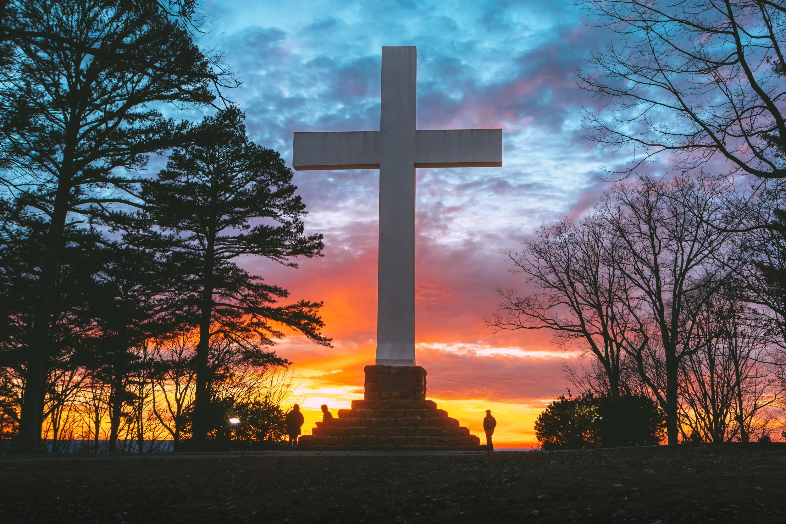 Large white cross monument on stepped base at sunset, silhouetted trees and people nearby, colorful sky with orange, pink, and blue clouds.