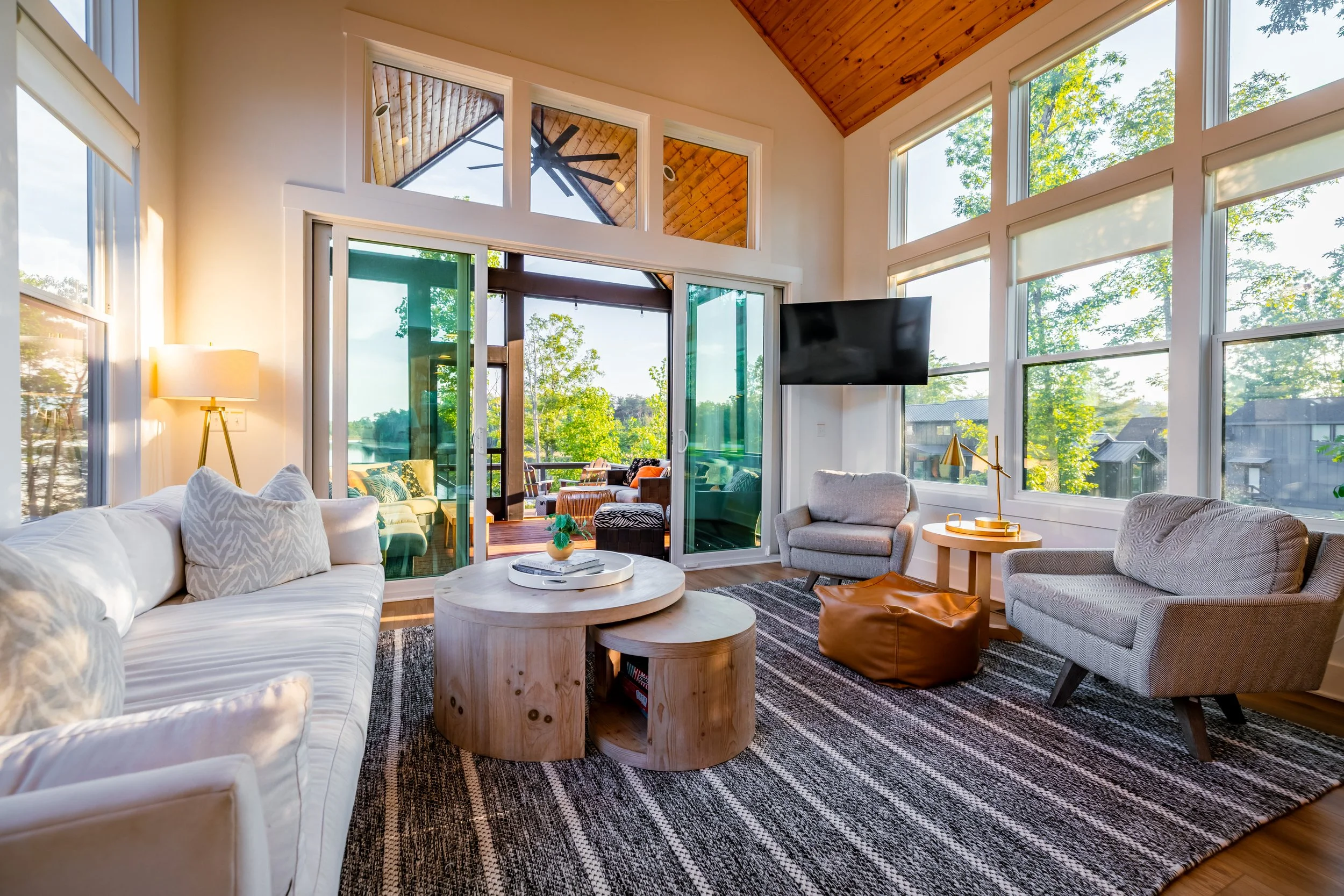 Bright living room with large windows, a white sofa, two gray armchairs, a wooden side table with a gold lamp, a black flat-screen TV, a black and gray striped rug, a round wooden coffee table, and a view of a deck with outdoor furniture and green tr