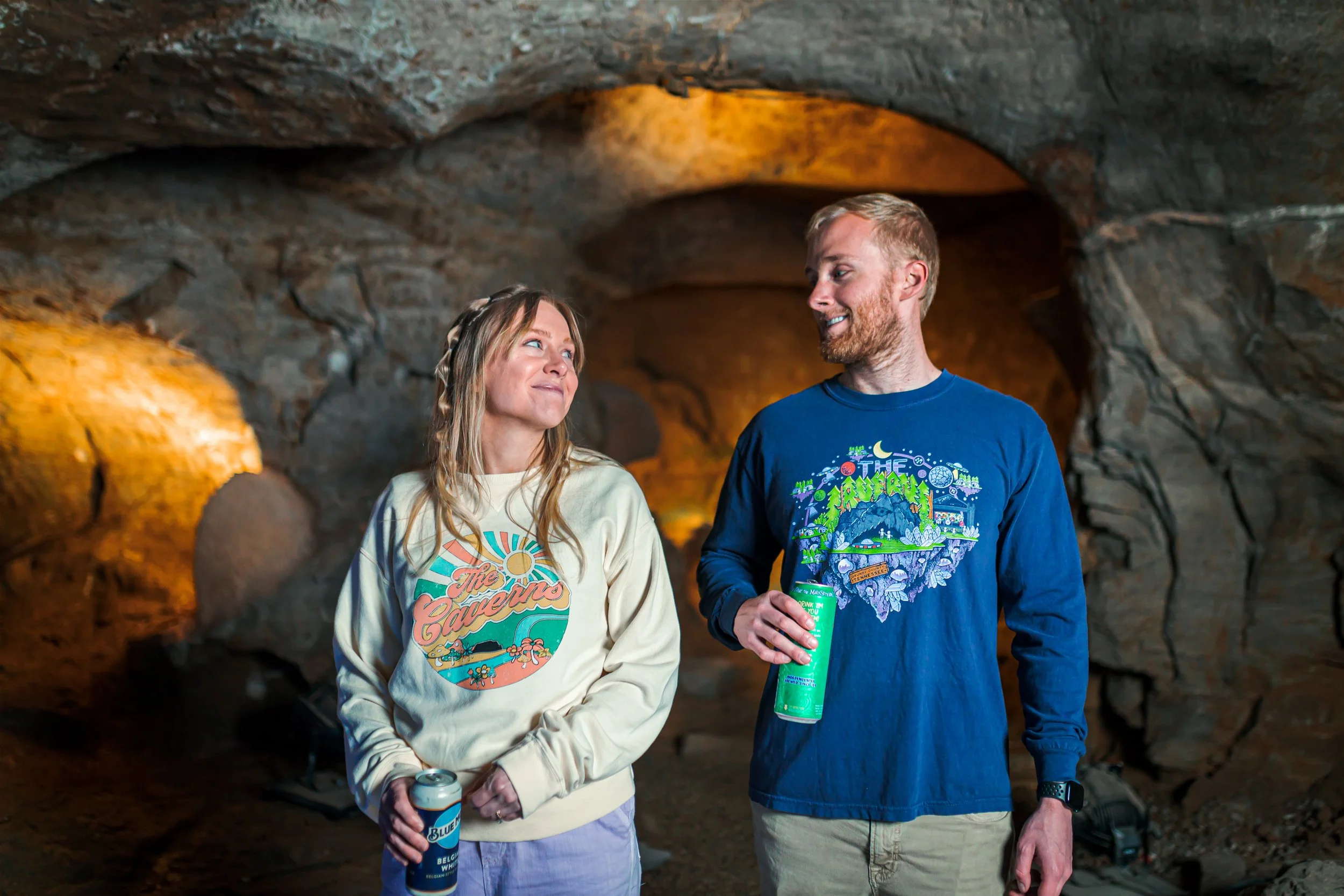 A woman and man stand inside a cave, smiling and holding drinks, with the woman wearing a cream sweatshirt and the man in a blue graphic shirt.