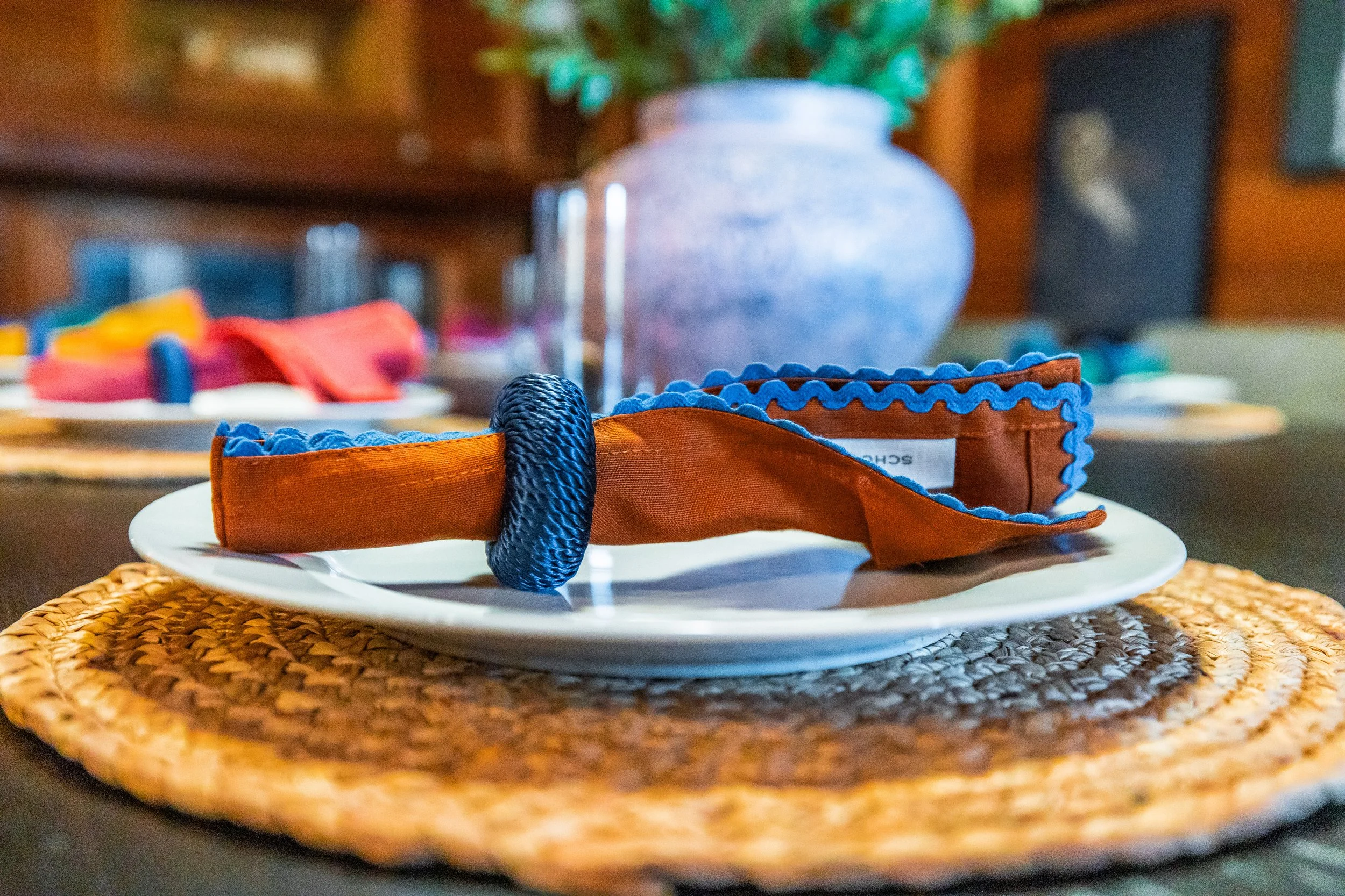 Close-up of a headband with orange fabric, blue elastic, and black braided ring, placed on a white plate on a woven placemat, with blurred background of a dining table and wooden wall.