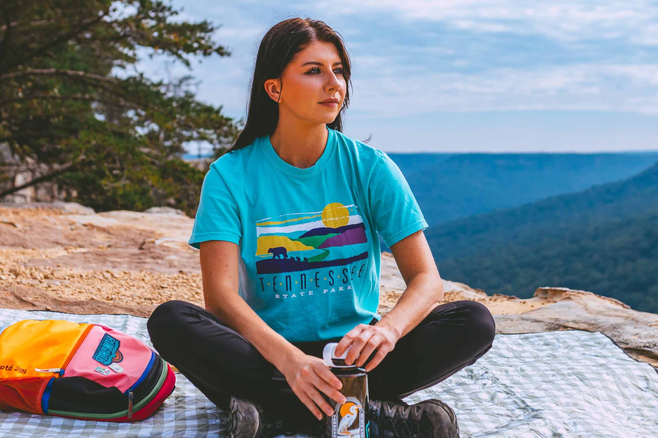 A woman sitting cross-legged on a blanket outdoors with a scenic mountain view, holding a drink, wearing a teal T-shirt with 'Tennessee State Park' printed on it, and a colorful backpack beside her.
