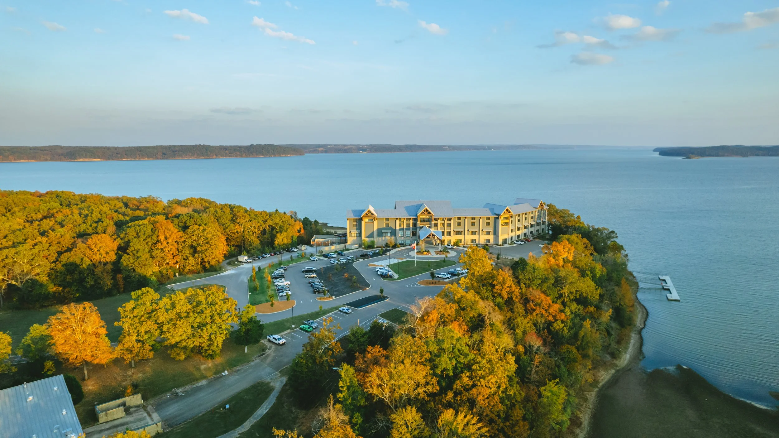 An aerial view of a large hotel on a cliff overlooking a body of water, with a parking lot and trees showing fall colors.
