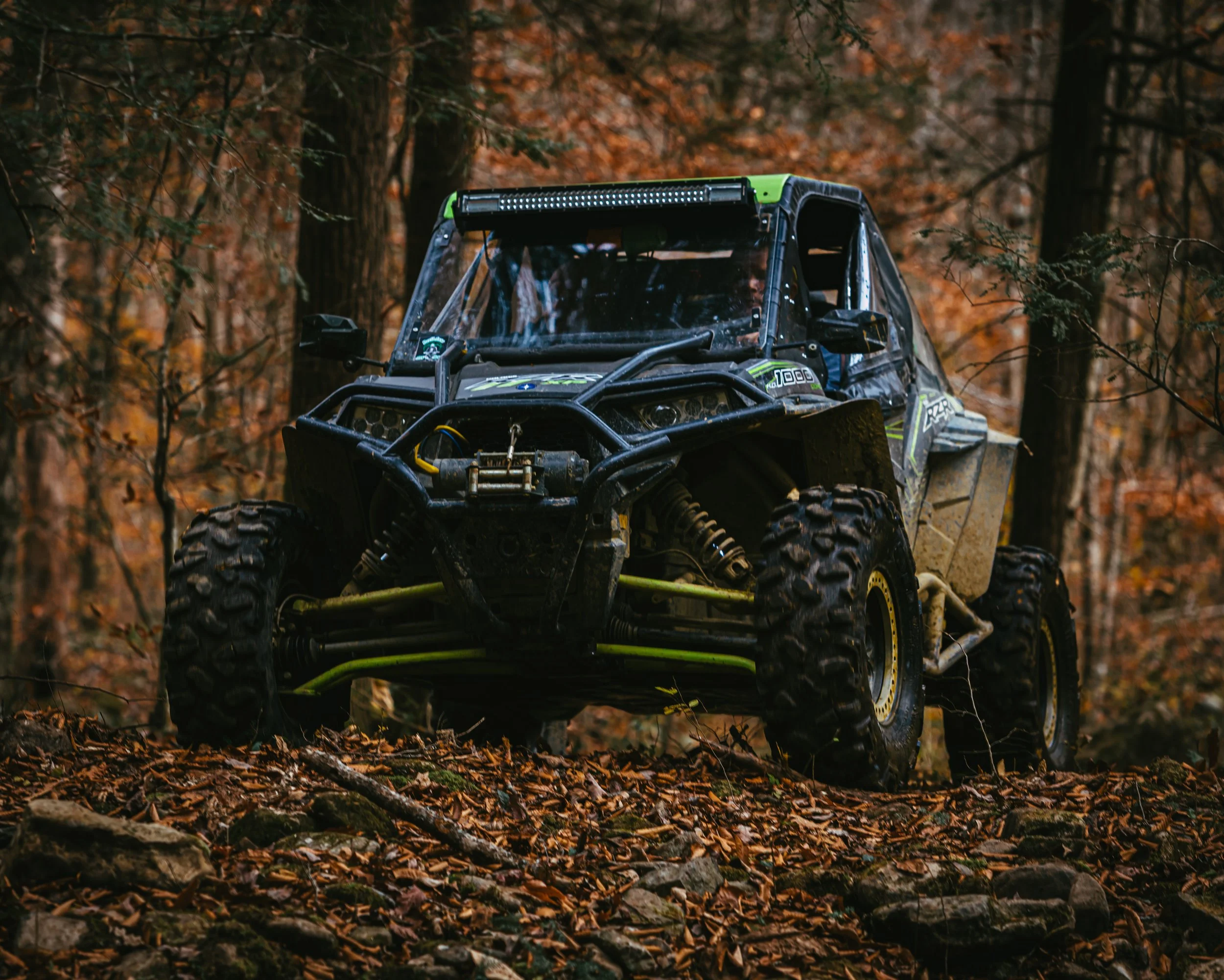 An off-road vehicle driving through a wooded area with autumn leaves on the ground.
