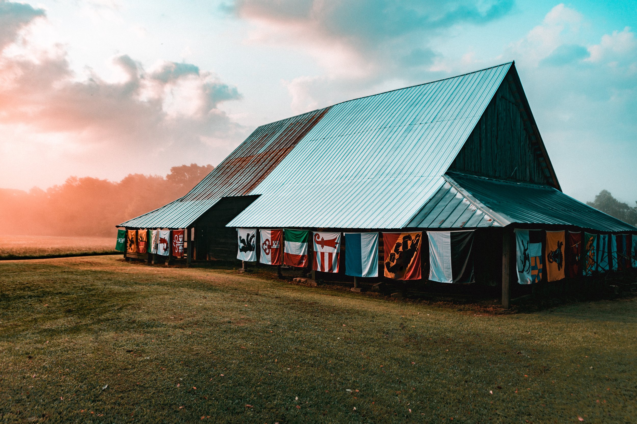 A rustic barn with a metal roof, decorated with various flags hanging on the side, outdoors during sunset with a grassy field and cloudy sky in the background.