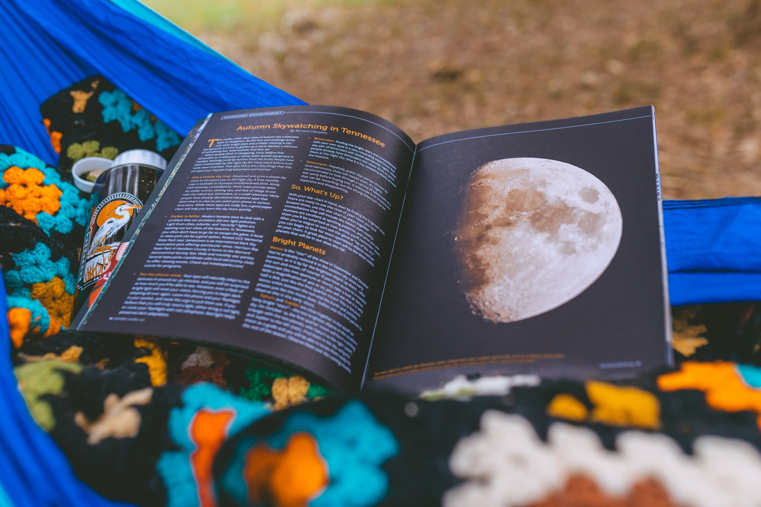 Open magazine with images and text about the Moon, resting on a blue hammock outdoors, next to a blanket with colorful crochet flowers and a can cooler with wildlife artwork.