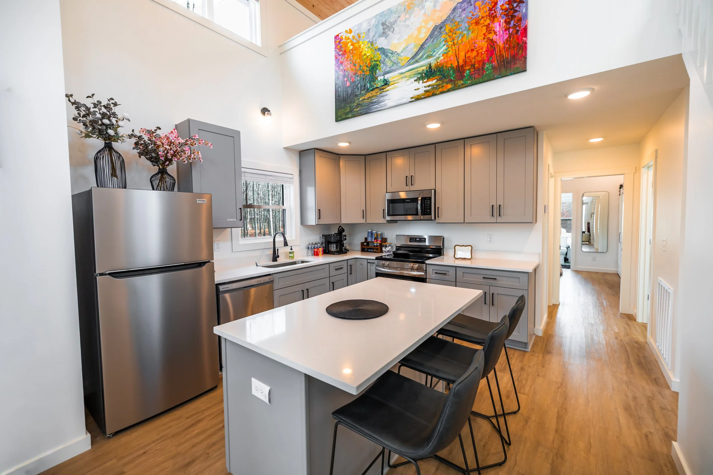 Modern kitchen with light gray cabinets, stainless steel refrigerator, microwave, and oven, white countertop, island with four black chairs, colorful landscape painting above cabinets, wooden floor, and a hallway leading to other rooms.