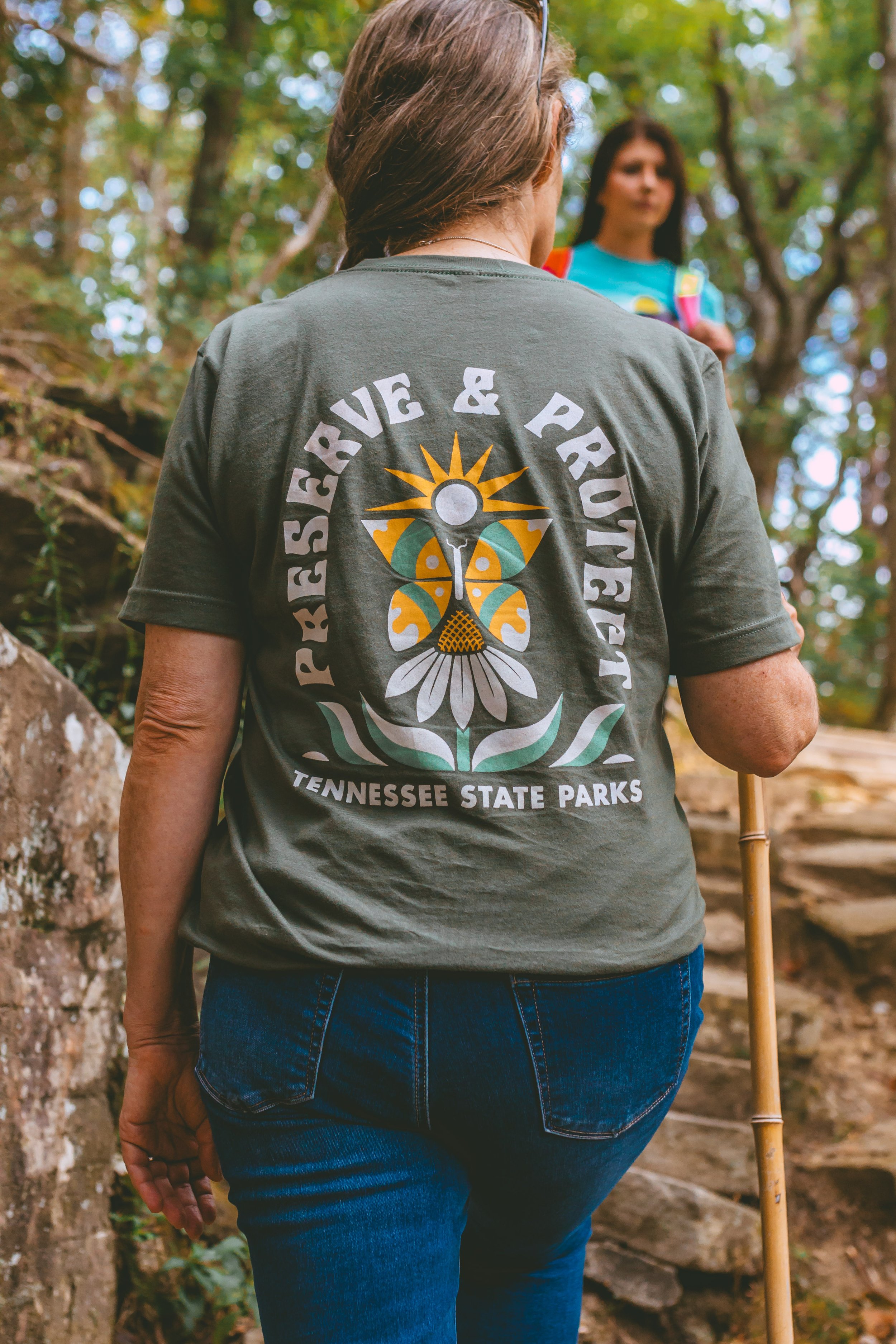 Back view of a woman wearing a Tennessee State Parks T-shirt with a butterfly and sun graphic, walking on a forest trail with another woman in the background.