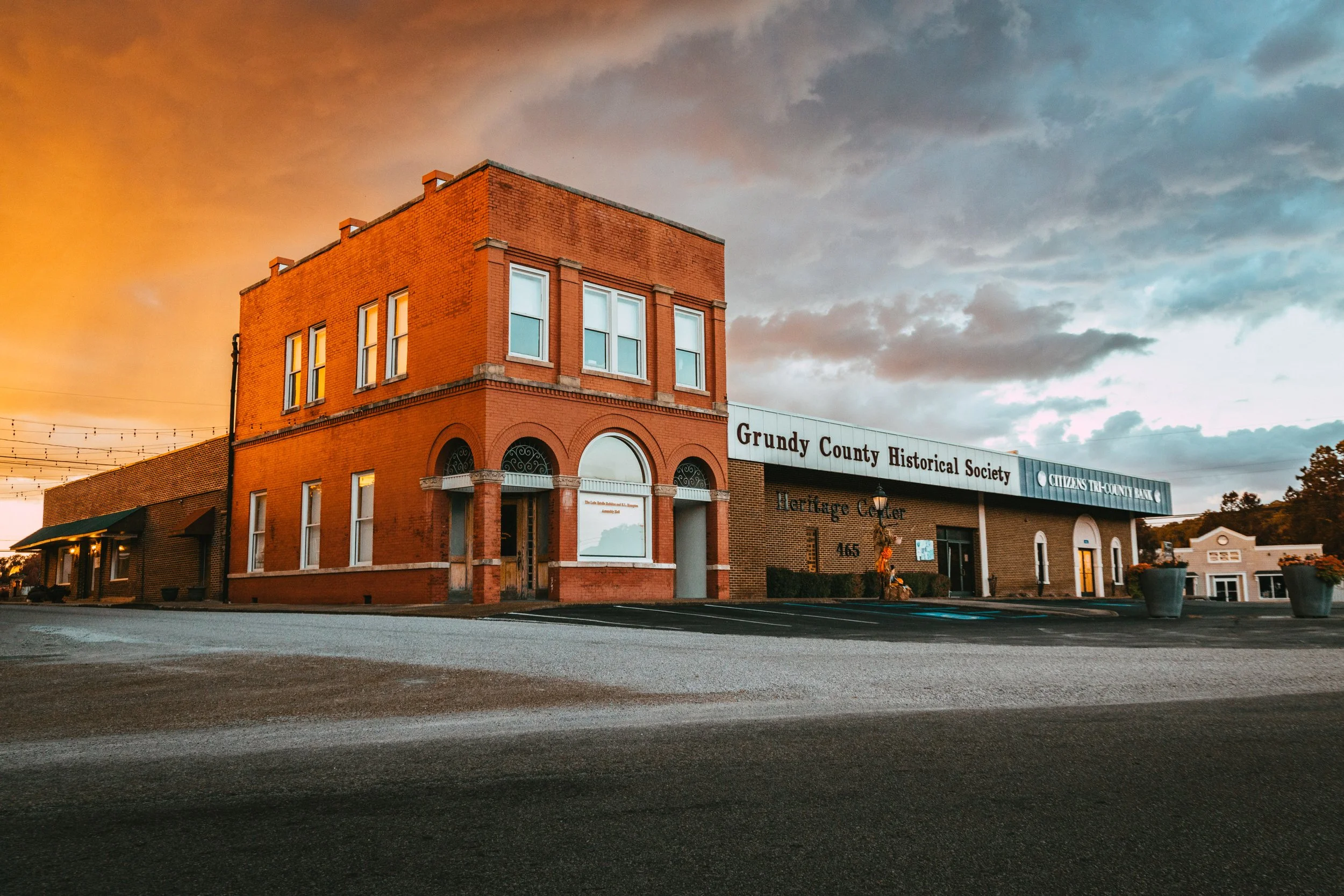 A brick building with a sign reading 'Gundy County Historical Society' and 'Heritage Center' is illuminated by a warm sunset sky. The building has large windows and an arched entrance.
