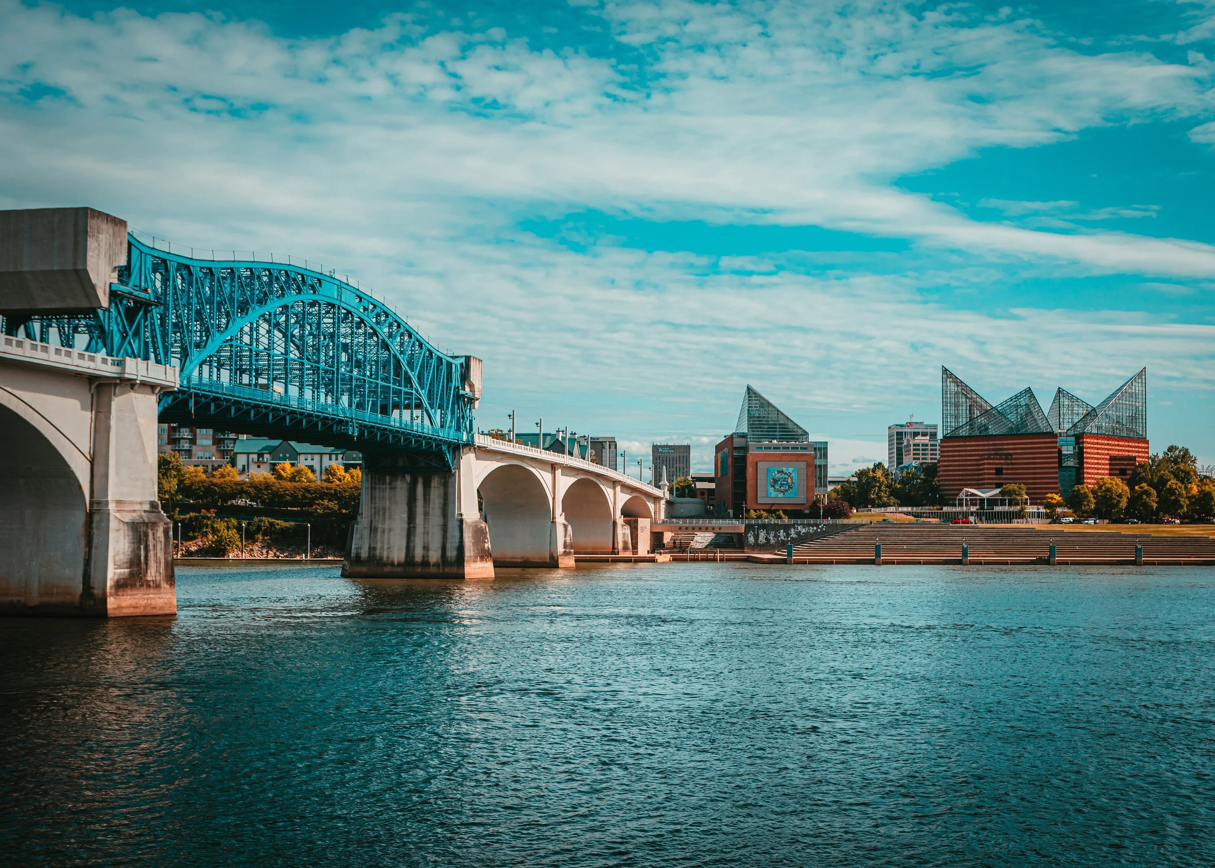 A city skyline with a blue bridge over a body of water, modern buildings including a red brick structure and glass skyscrapers, under a partly cloudy sky.