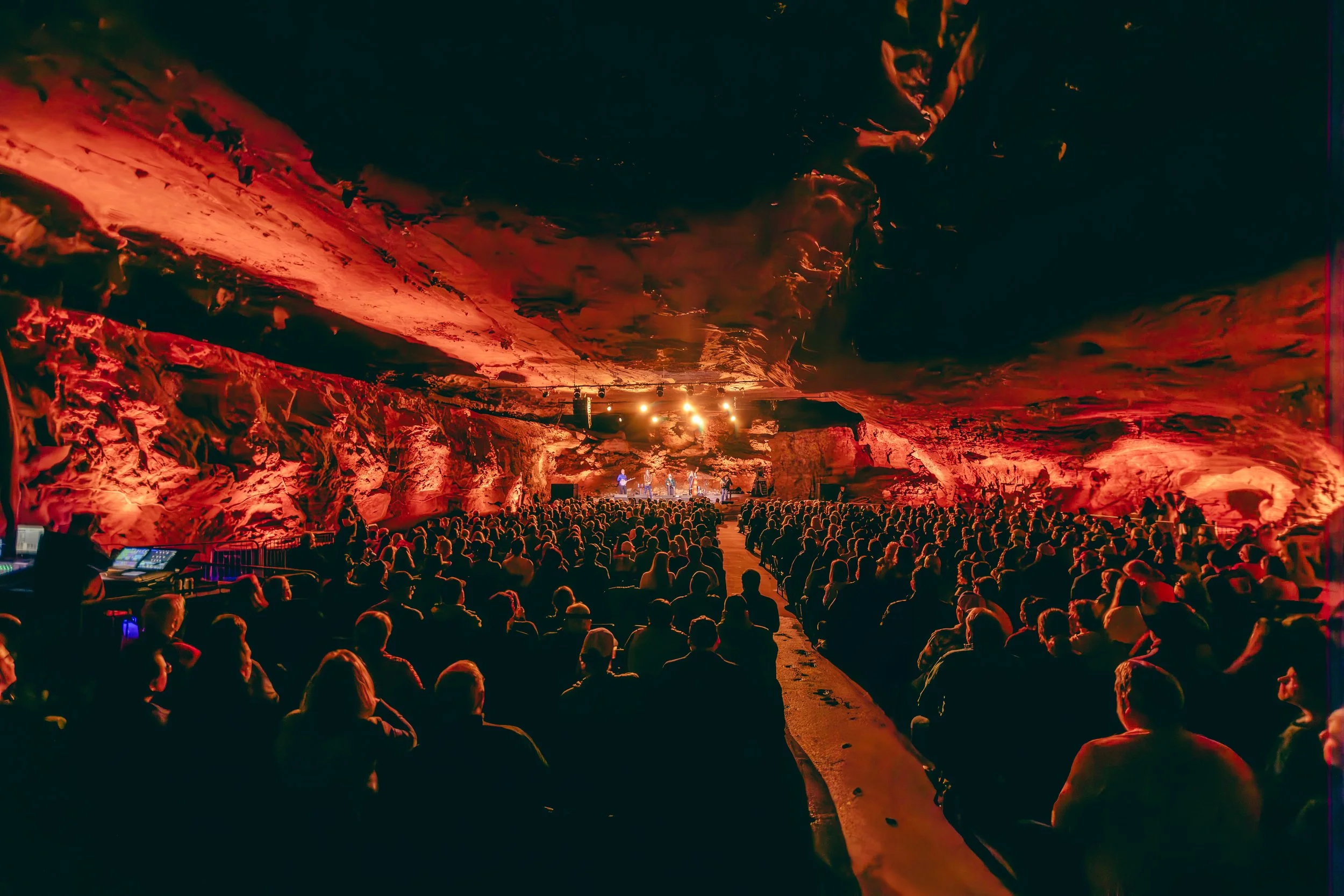 An underground concert with a large audience facing a stage, illuminated with red and orange lighting, surrounded by rock walls and ceiling.