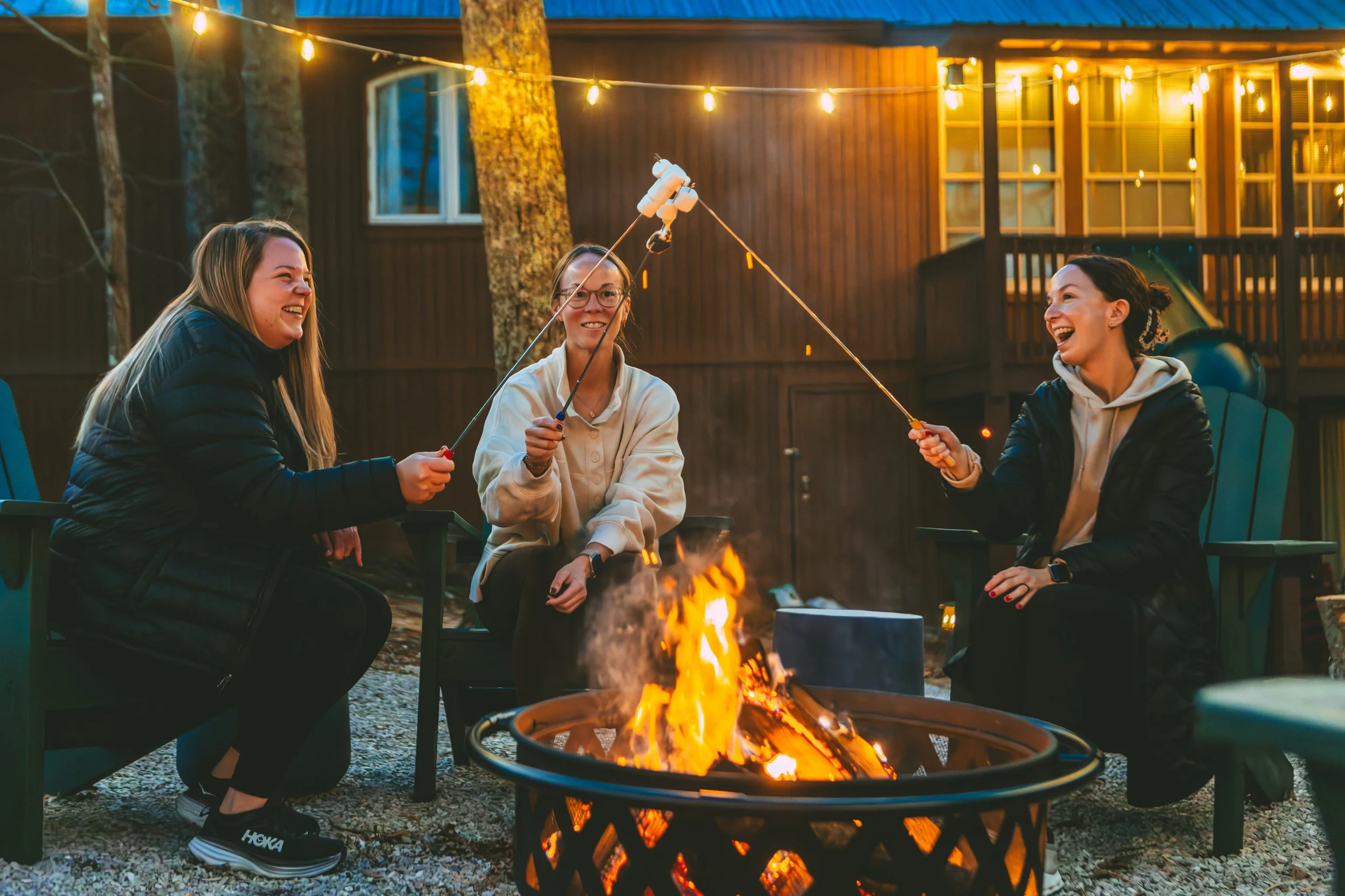 Three women sitting around a fire pit outdoors, roasting marshmallows on sticks, smiling and laughing, with string lights hanging overhead at night.