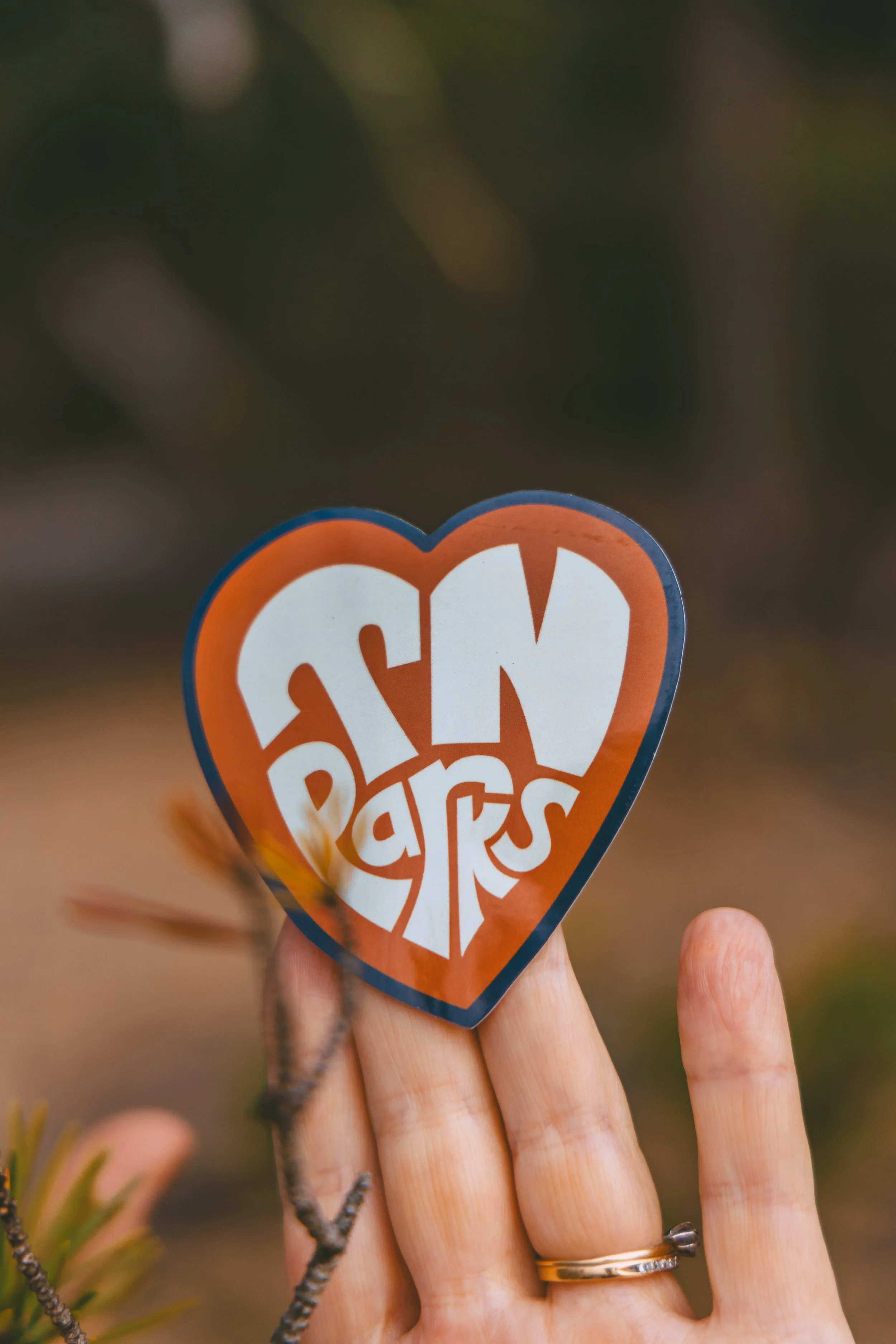 Hand holding a heart-shaped sticker with the words 'Stay Weird' in bold white letters on an orange background.
