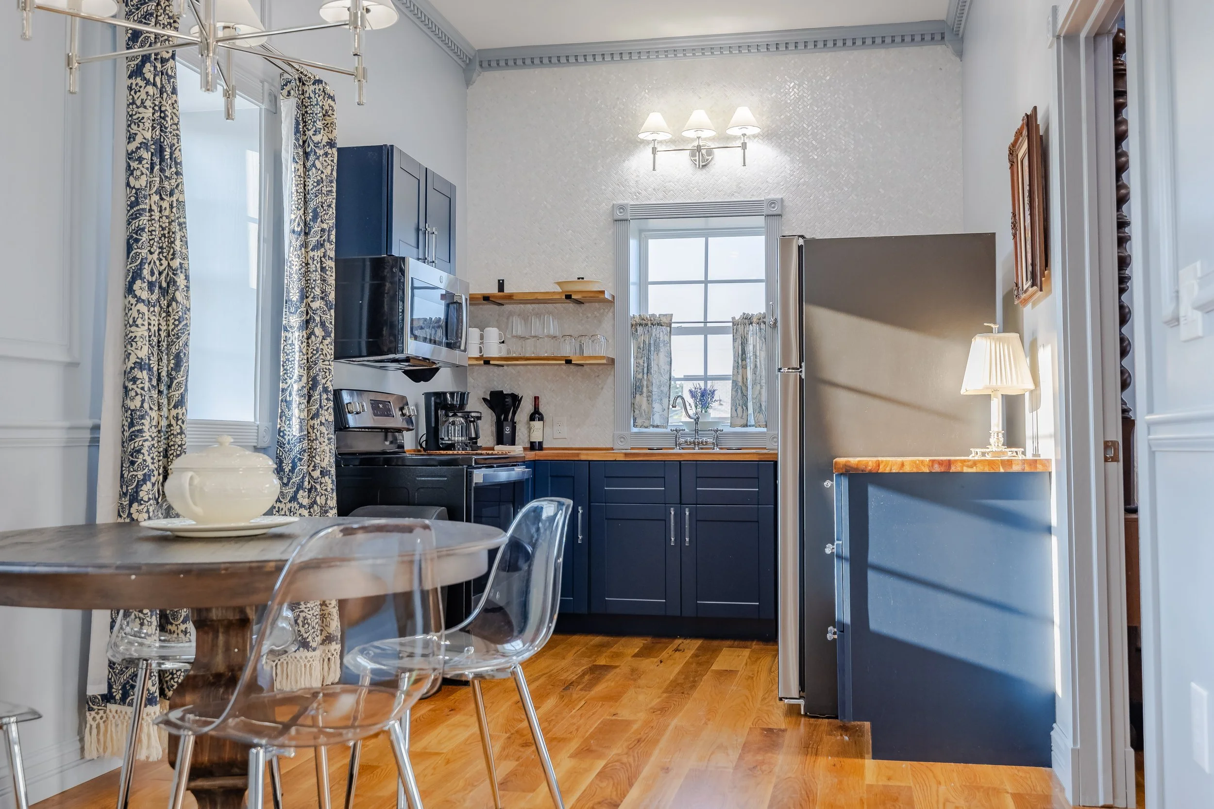 A cozy kitchen with blue cabinets, wooden countertops, and white textured walls. There are open shelves with glassware, a window with patterned curtains, and a refrigerator. A small table with clear plastic chairs and a ceramic teapot is in the foreg