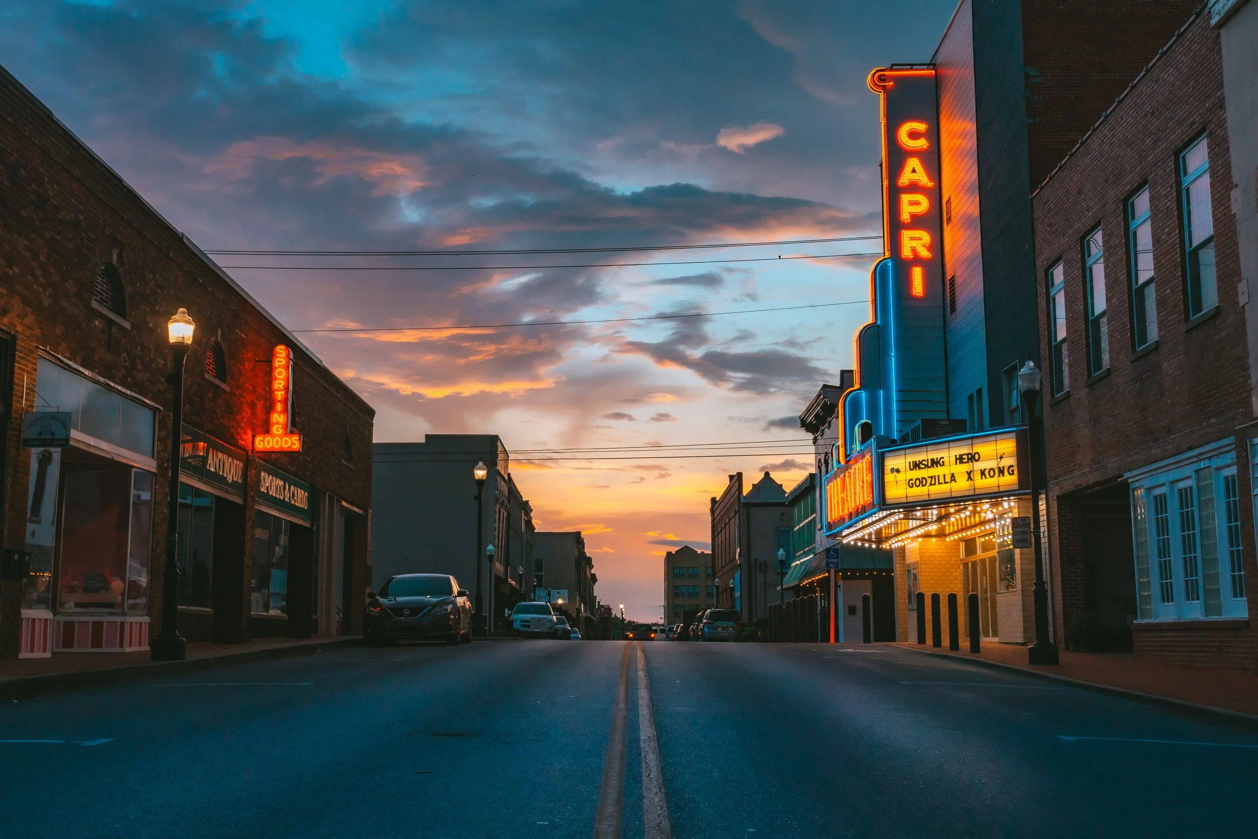 Empty street at sunset with buildings featuring neon signs and a movie theater marquee.