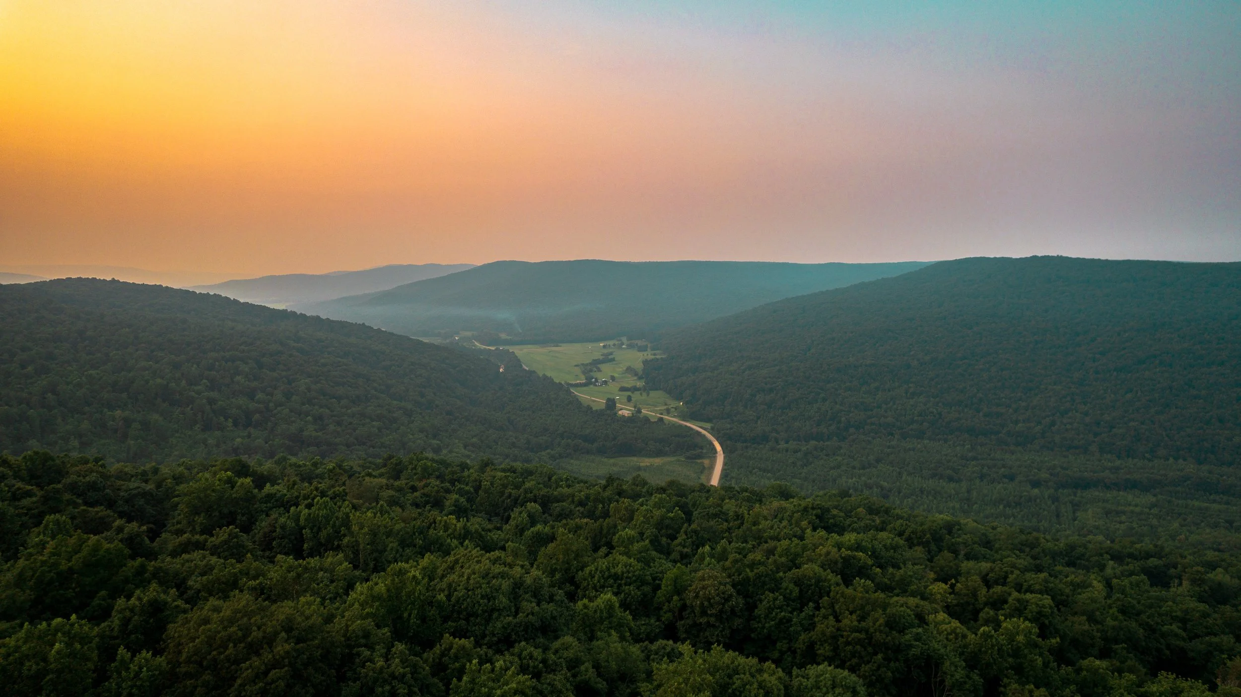Landscape of green forested mountains during sunset with a winding road through the valley.
