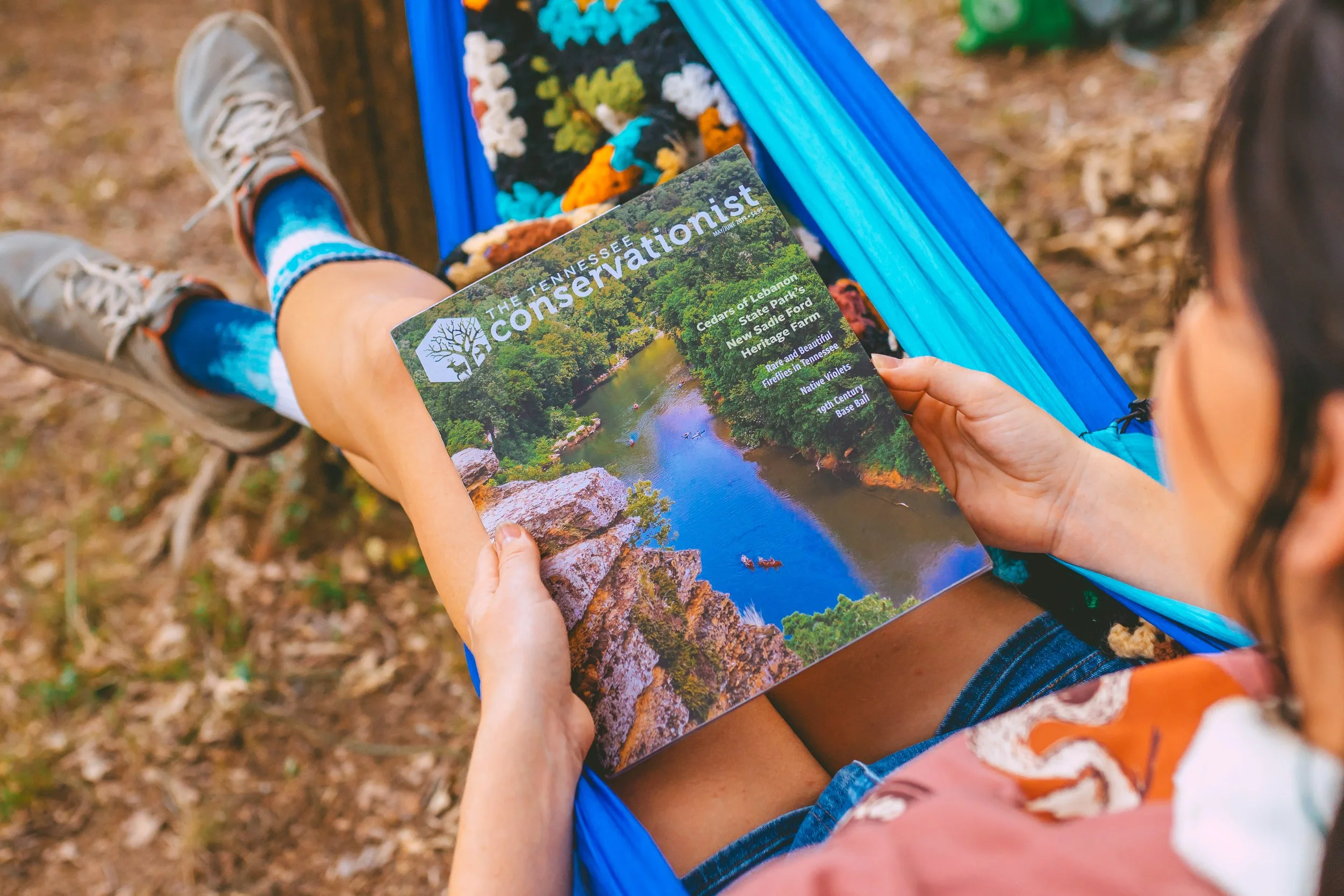 A person sitting in a blue hammock outdoors, reading a magazine titled "The Tennessee Conservationist" with a cover image of a river and trees.
