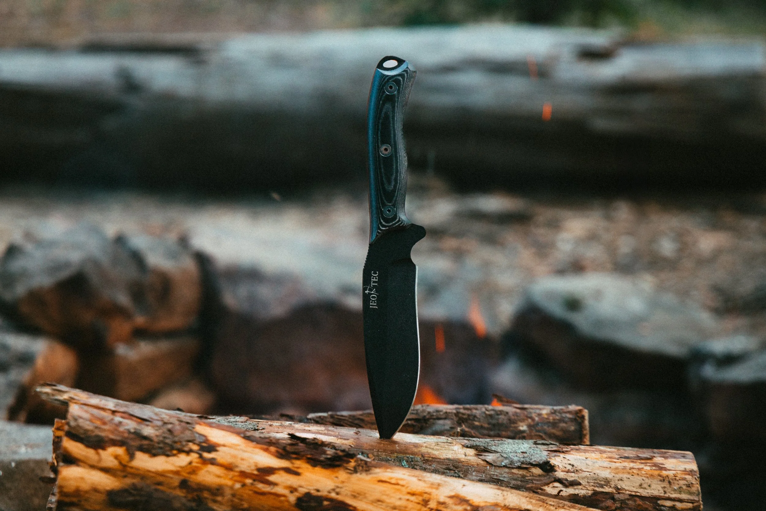 A black hunting knife with a patterned handle stuck into a piece of wood outdoors, with logs and rocks in the background.