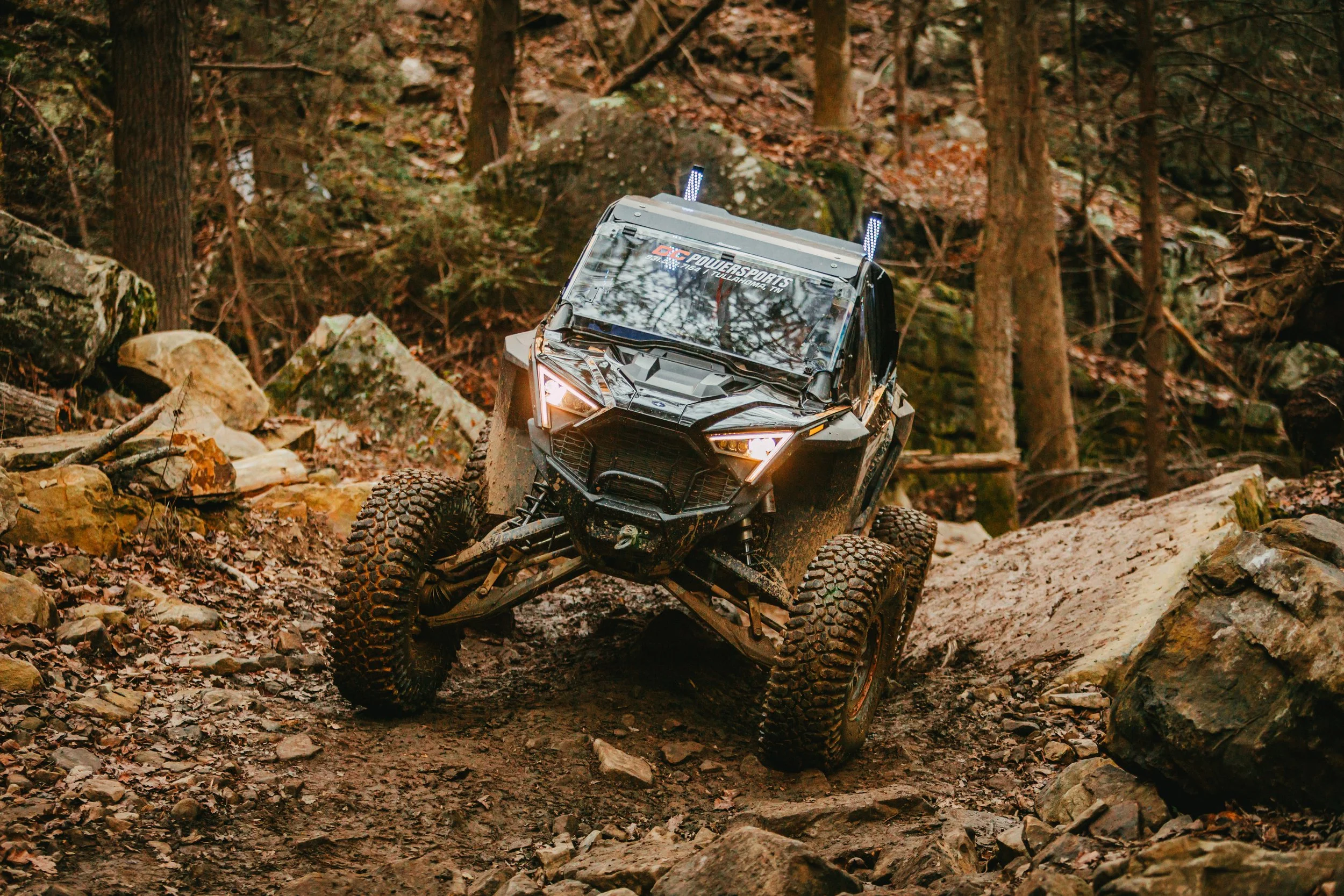 An off-road vehicle navigating a rocky forest trail with trees and stones around.