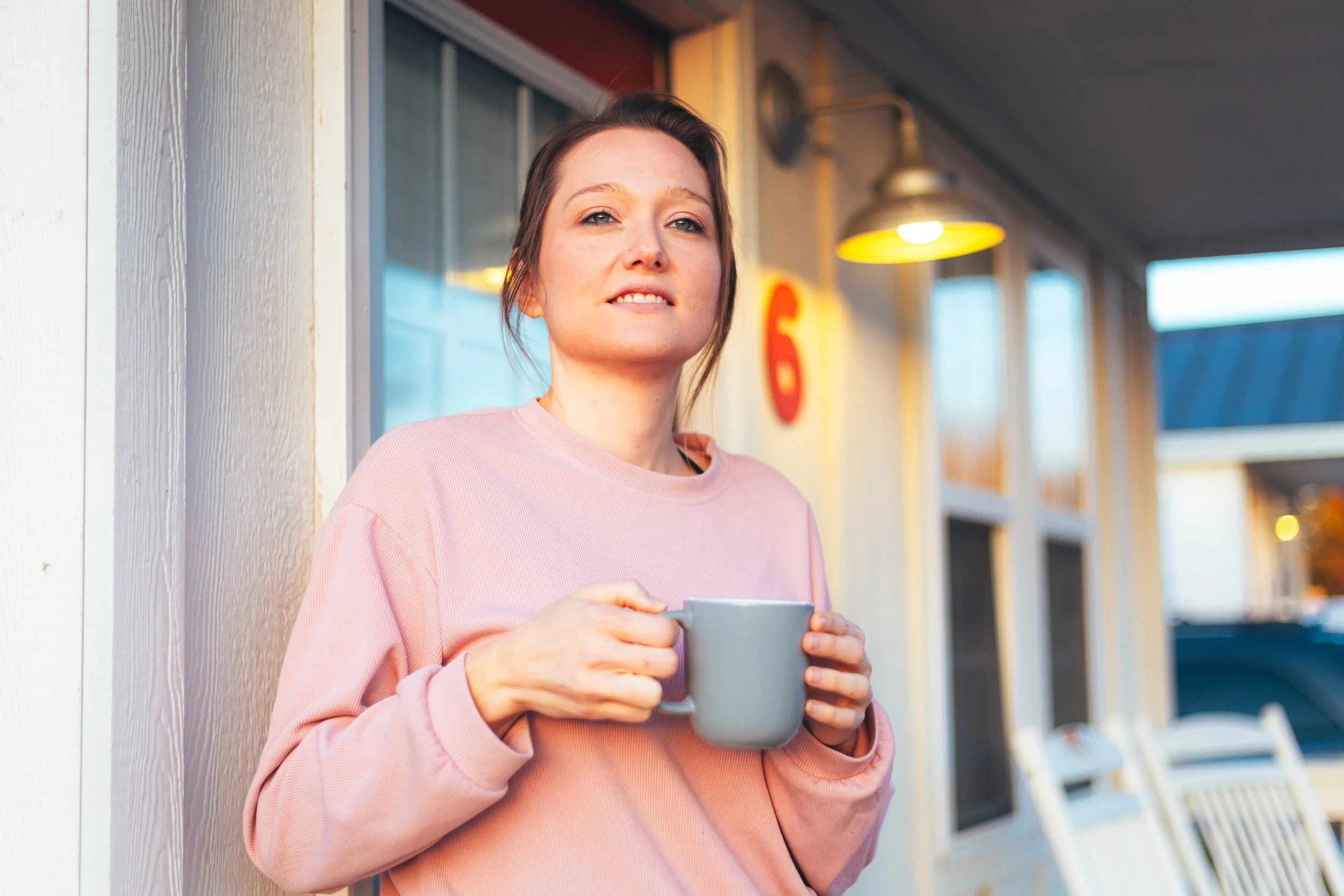 A woman with brown hair in a pink sweatshirt holding a gray mug, standing outside a building with large windows and a white wall, during sunset.