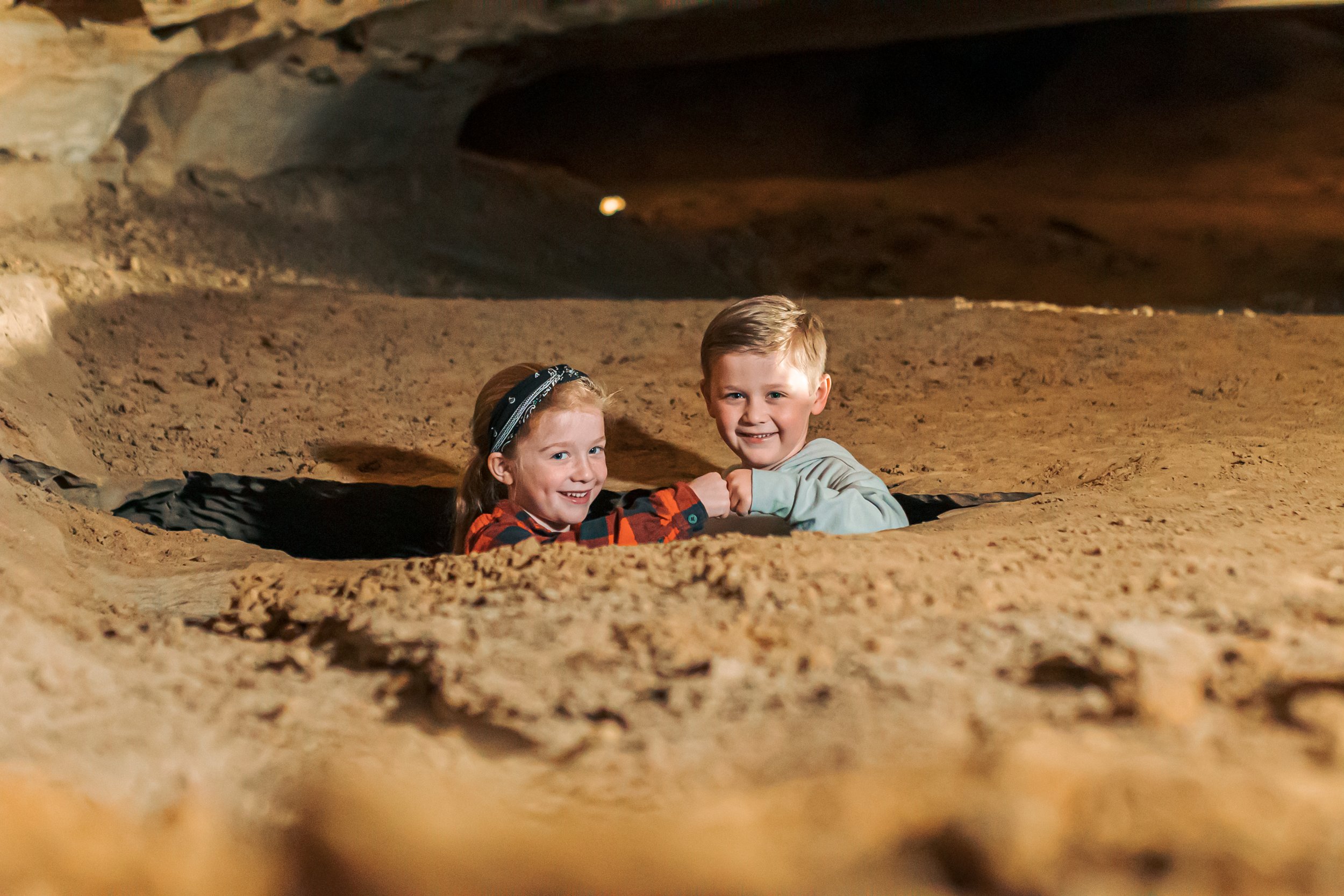 Two children, a girl and a boy, smiling and holding hands while sitting in a hole in sandy terrain inside a cave or underground area.