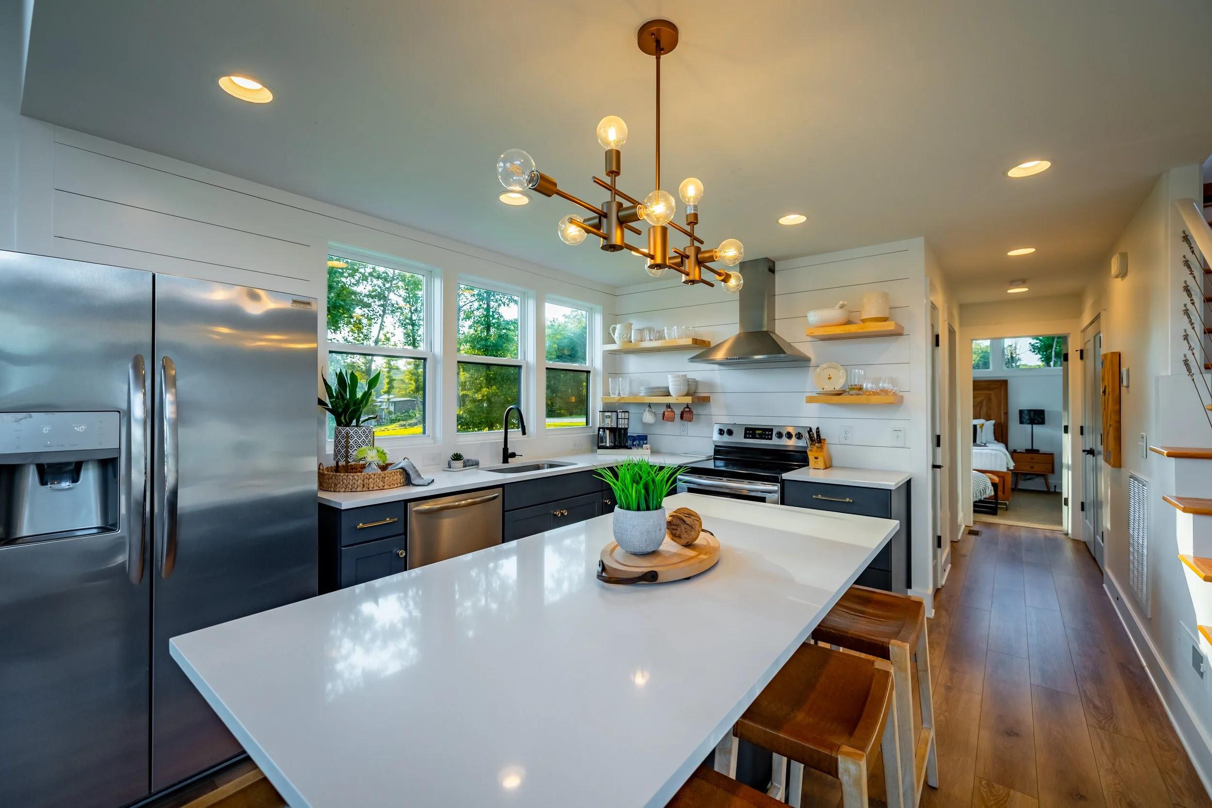 Bright modern kitchen with white island, dark cabinets, stainless steel appliances, open shelving, and large windows, flowing into a bedroom.