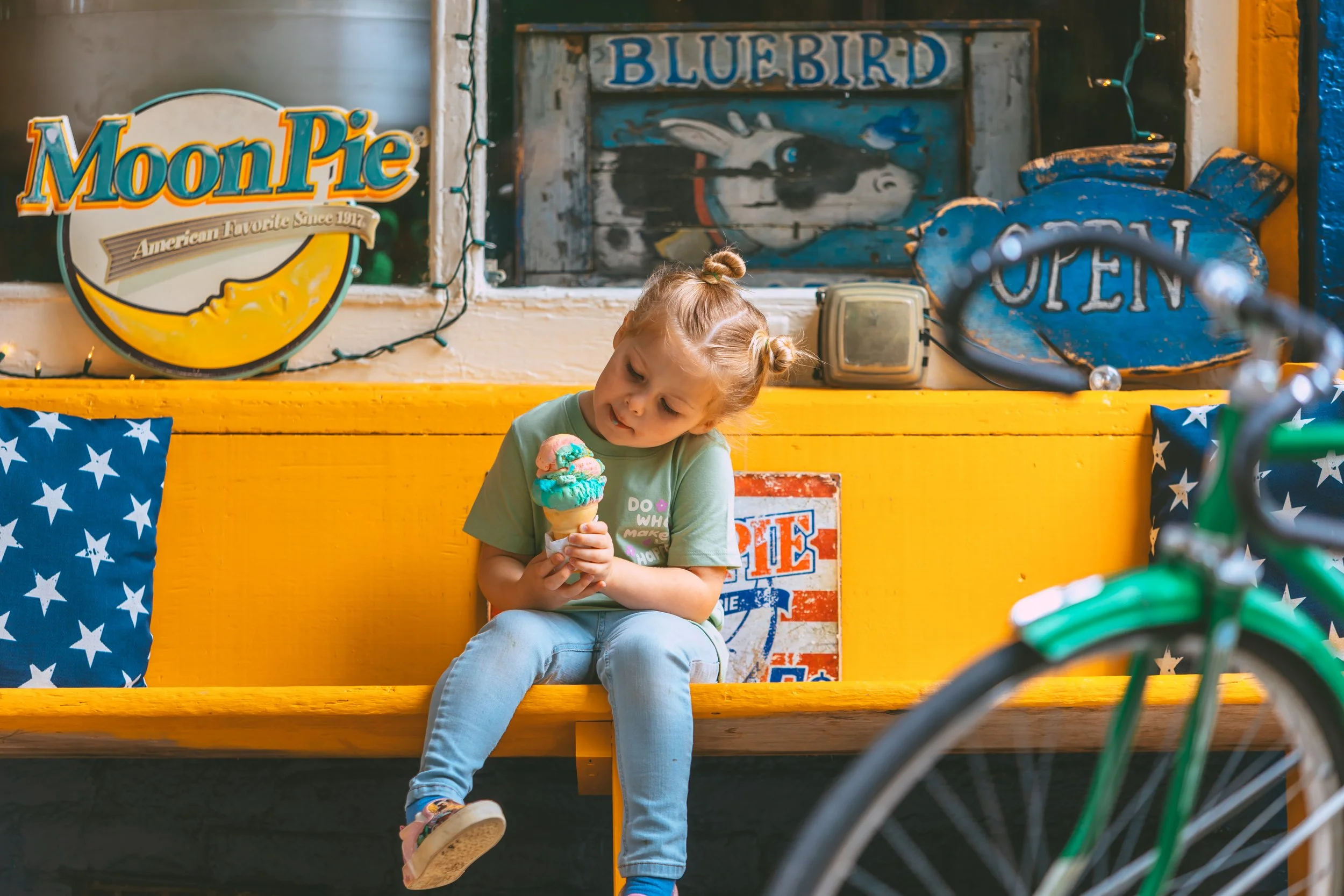 A young girl with two buns in her hair, sitting on a yellow wooden bench, holding and looking at a colorful ice cream cone inside an ice cream shop with vintage signs and a blue pillow with white stars behind her.