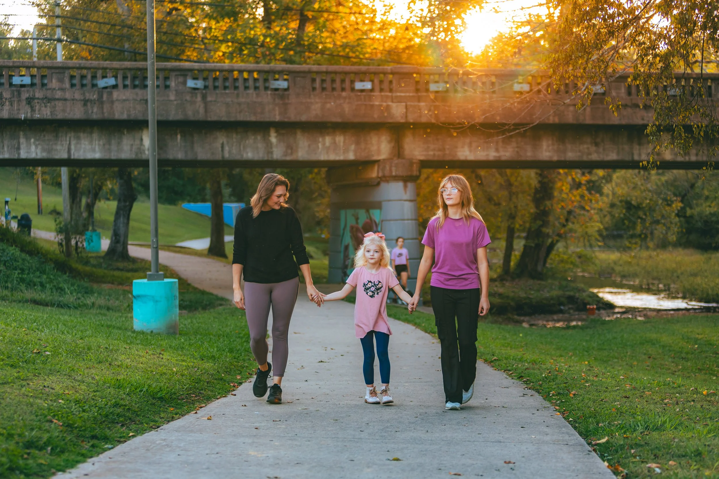 Three people, two women and a young girl, walk hand-in-hand along a paved path under a bridge in a park during sunset. The woman on the left wears a black top and gray leggings, the girl in the middle wears a pink dress with a heart design, and the w