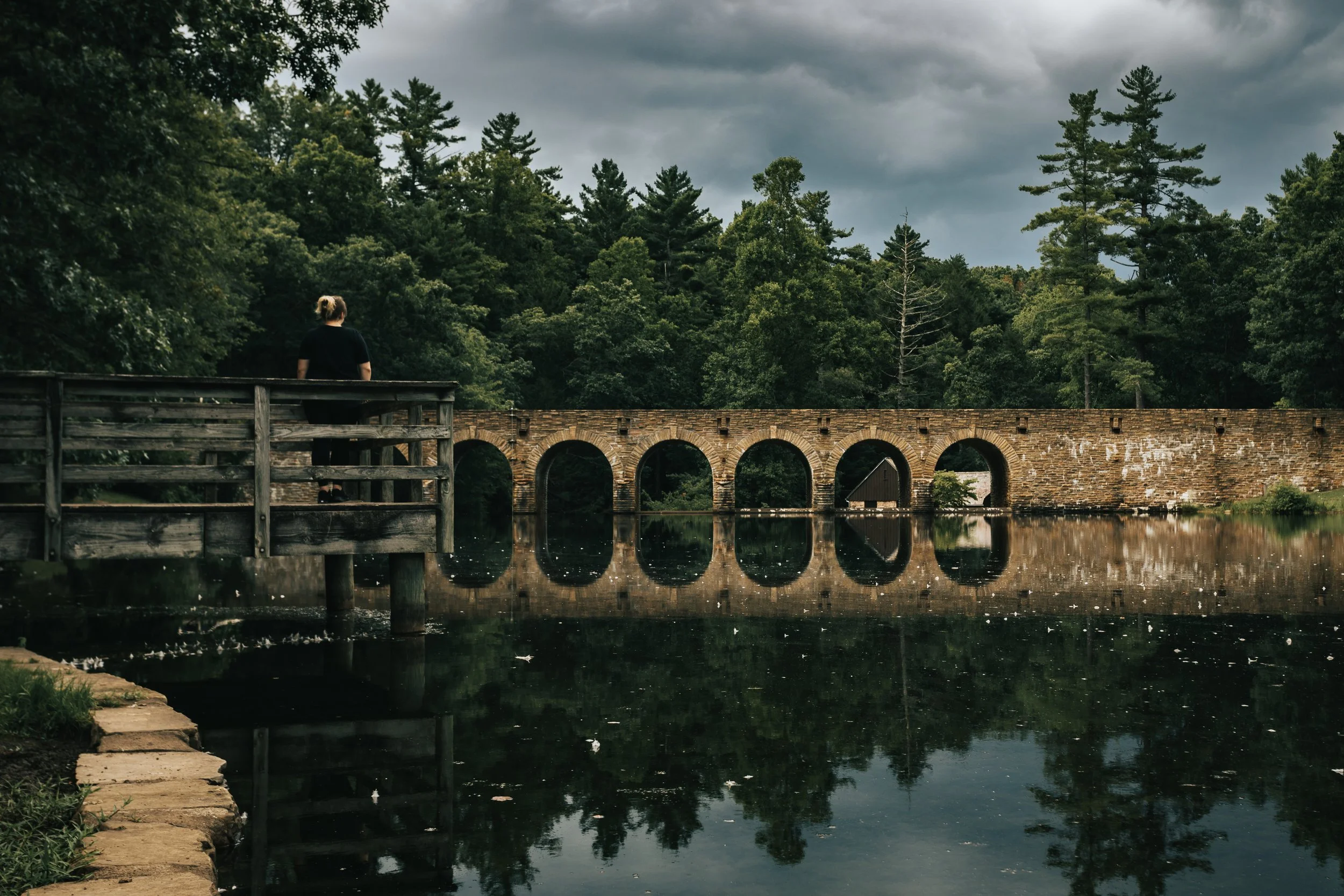 A person standing on a wooden dock looking at a stone arch bridge over a calm body of water, surrounded by trees and an overcast sky.