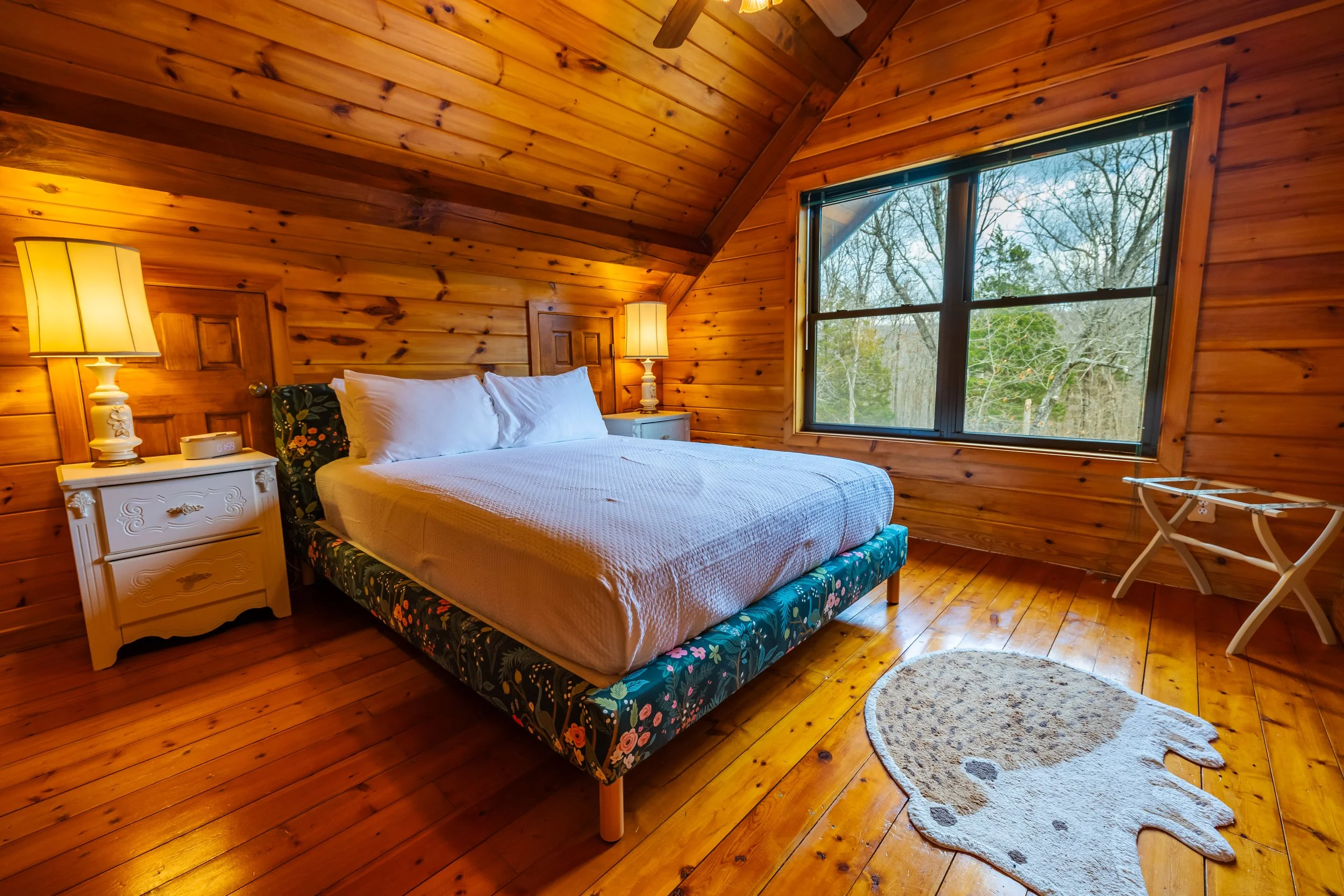A cozy bedroom with wooden walls and ceiling, a large bed with white pillows and a floral bed frame, two white nightstands with lamps, a window showing trees outside, a small rug shaped like a bear, and a folding luggage rack.