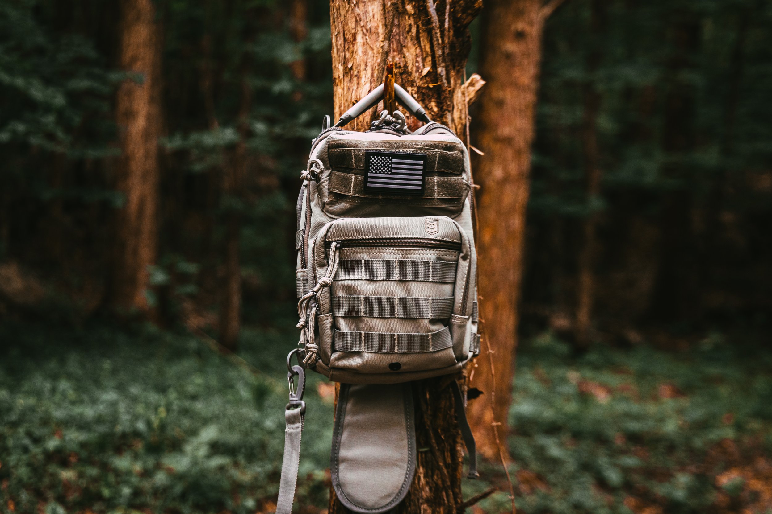A tactical backpack hanging on a tree in a forest, with an American flag patch on the top compartment.