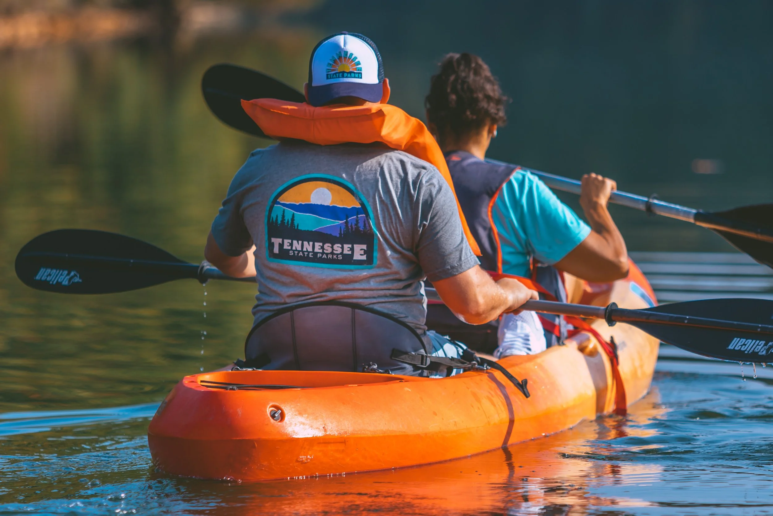 Two people kayaking on a lake, one wearing a Tennessee State Parks shirt and hat, the other in a blue shirt, paddling in an orange kayak.