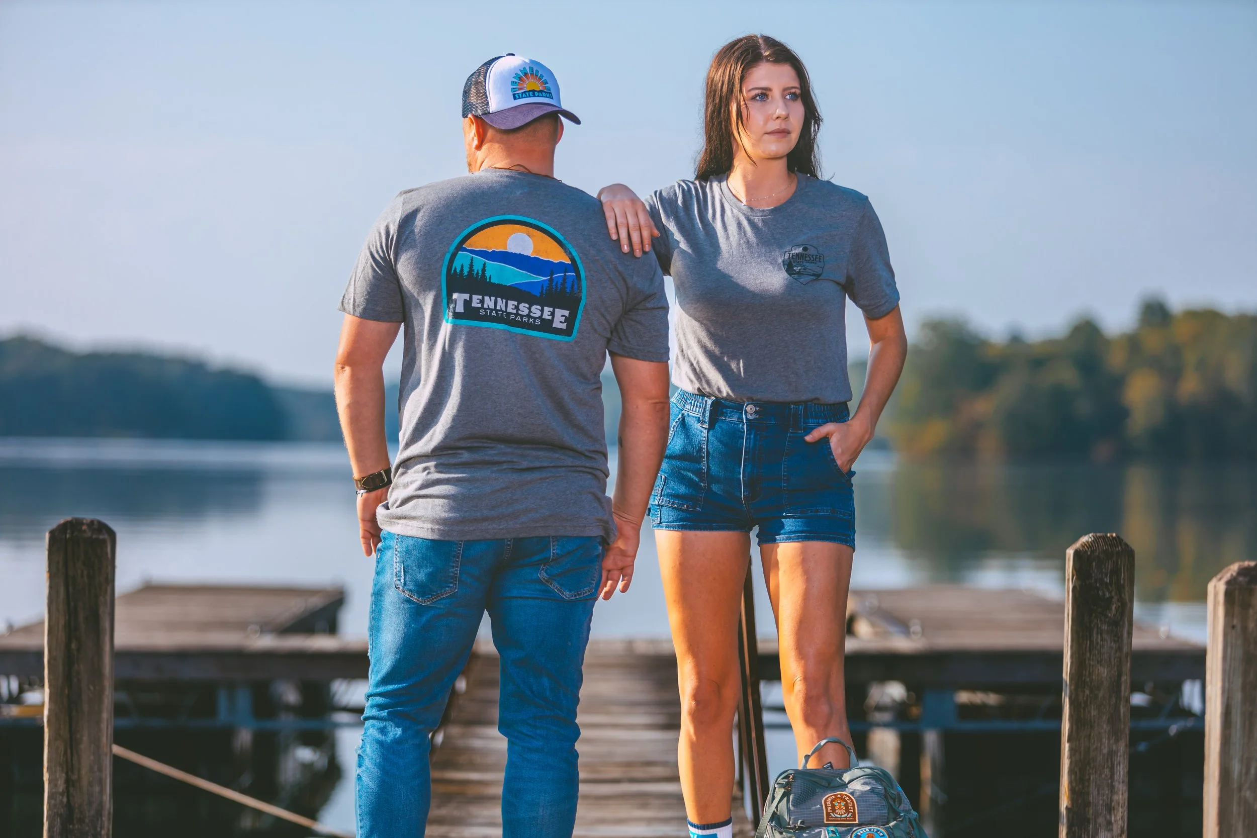 A man and woman standing on a wooden dock by a lake, with trees in the background. The man is facing away with a Tennessee State Parks shirt and cap. The woman is facing forward, wearing a gray T-shirt and denim shorts, with a backpack on the ground.