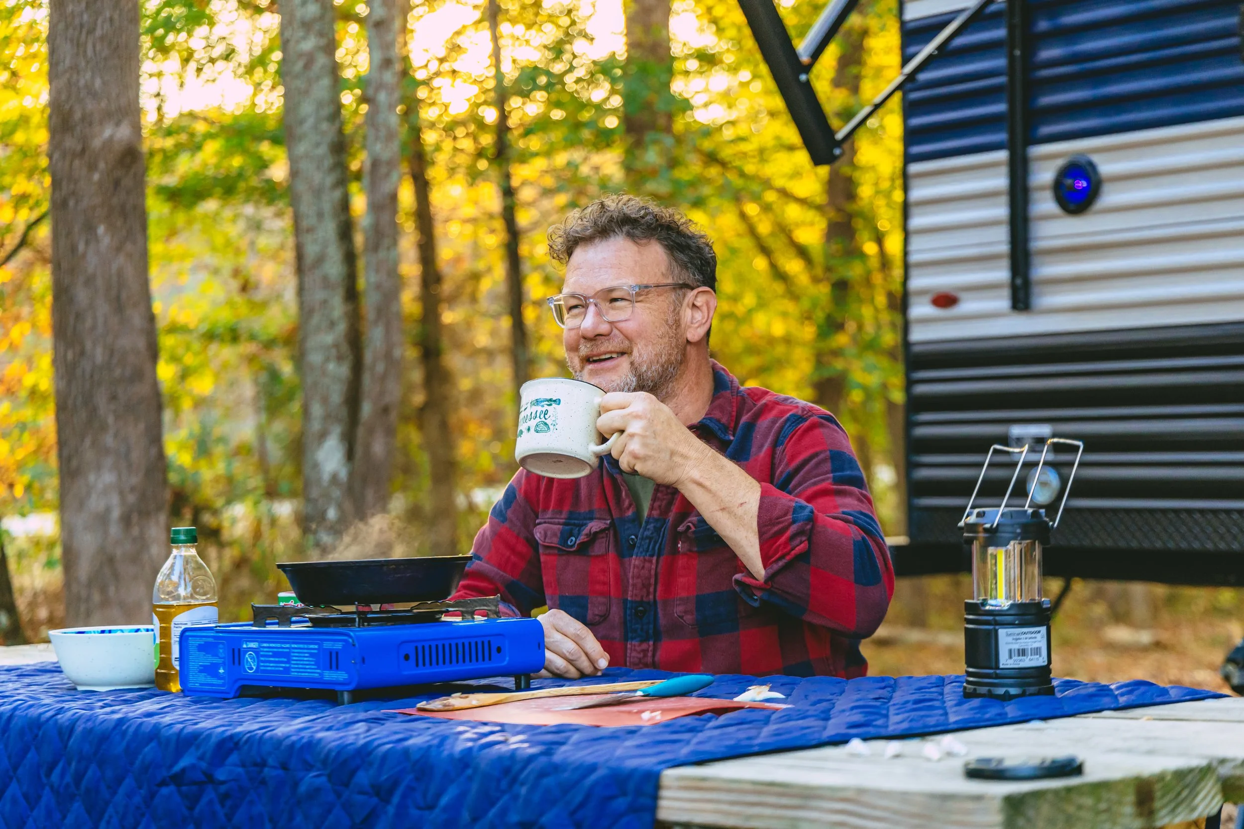 A man outdoors enjoying a camping meal, sitting at a table with a blue tablecloth, drinking from a mug, with a camping stove, bowl, and bottle on the table, and a trailer in the background during autumn.