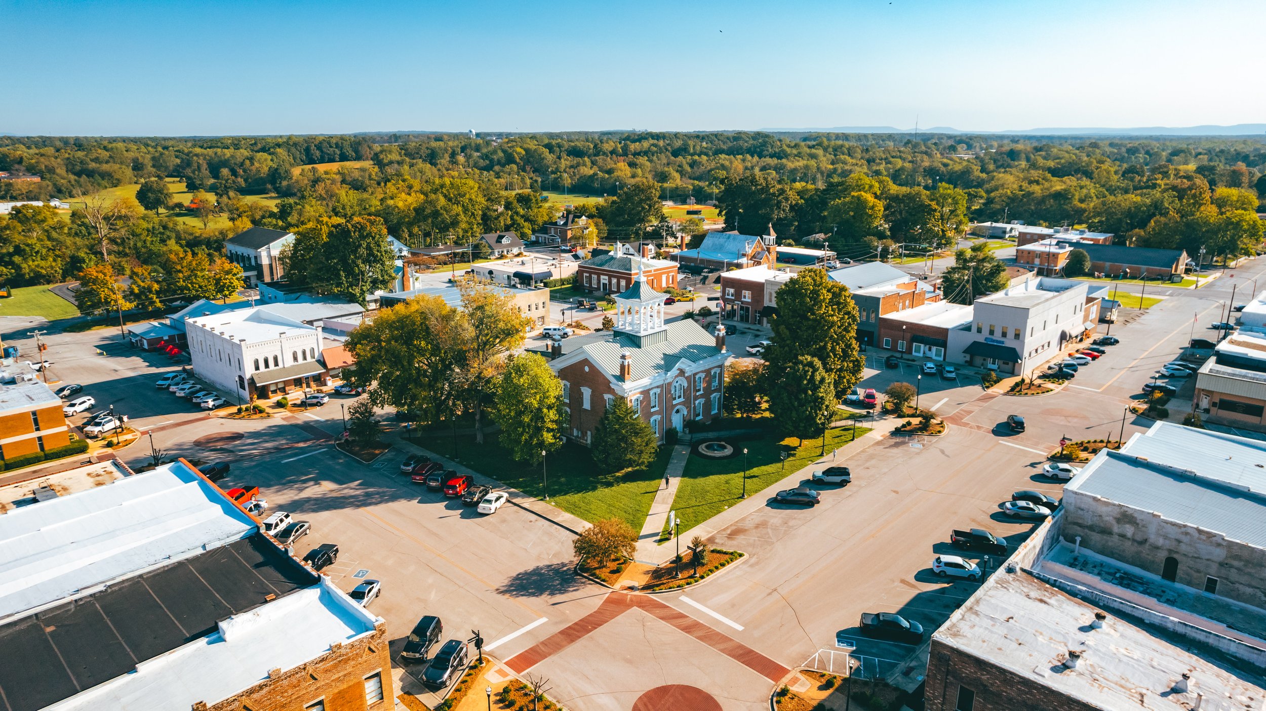 Bird's eye view of a small town square with a historic courthouse at the center, surrounded by parking lots and a mix of historic and modern buildings, green trees, and streets.
