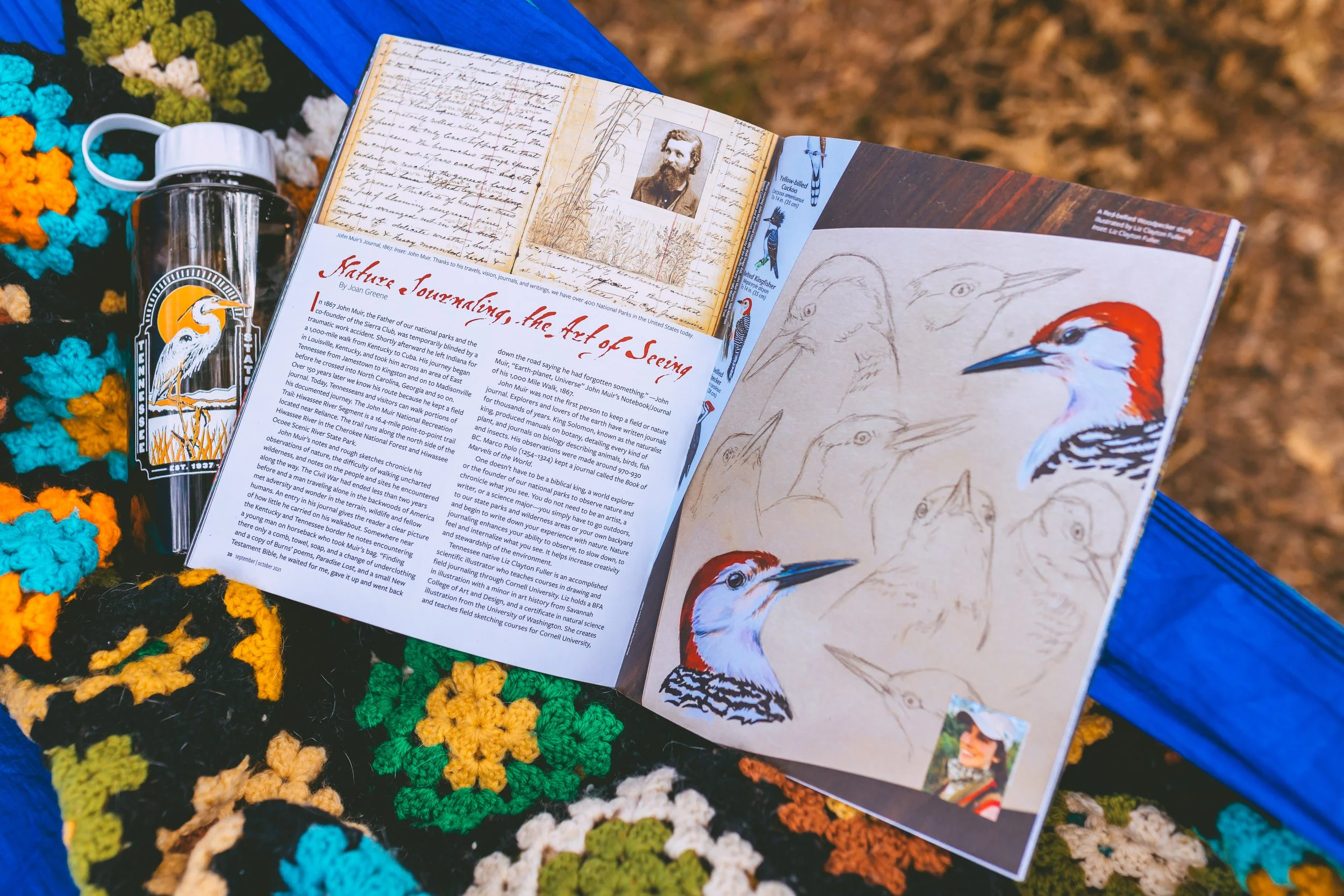 Open magazine on colorful crocheted blanket with a travel mug nearby. The magazine features illustrations and articles about nature journaling and birds, including sketches of woodpeckers and a photograph of a man with a beard.