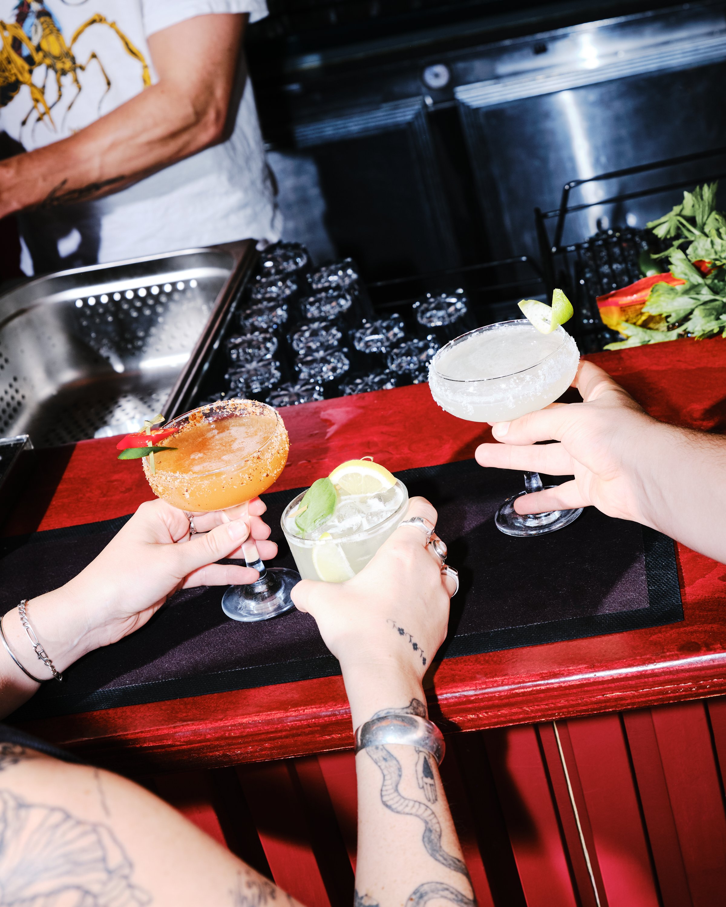Three people holding cocktails at a bar with a wood counter, fresh garnishes, and an ice bucket in the background.