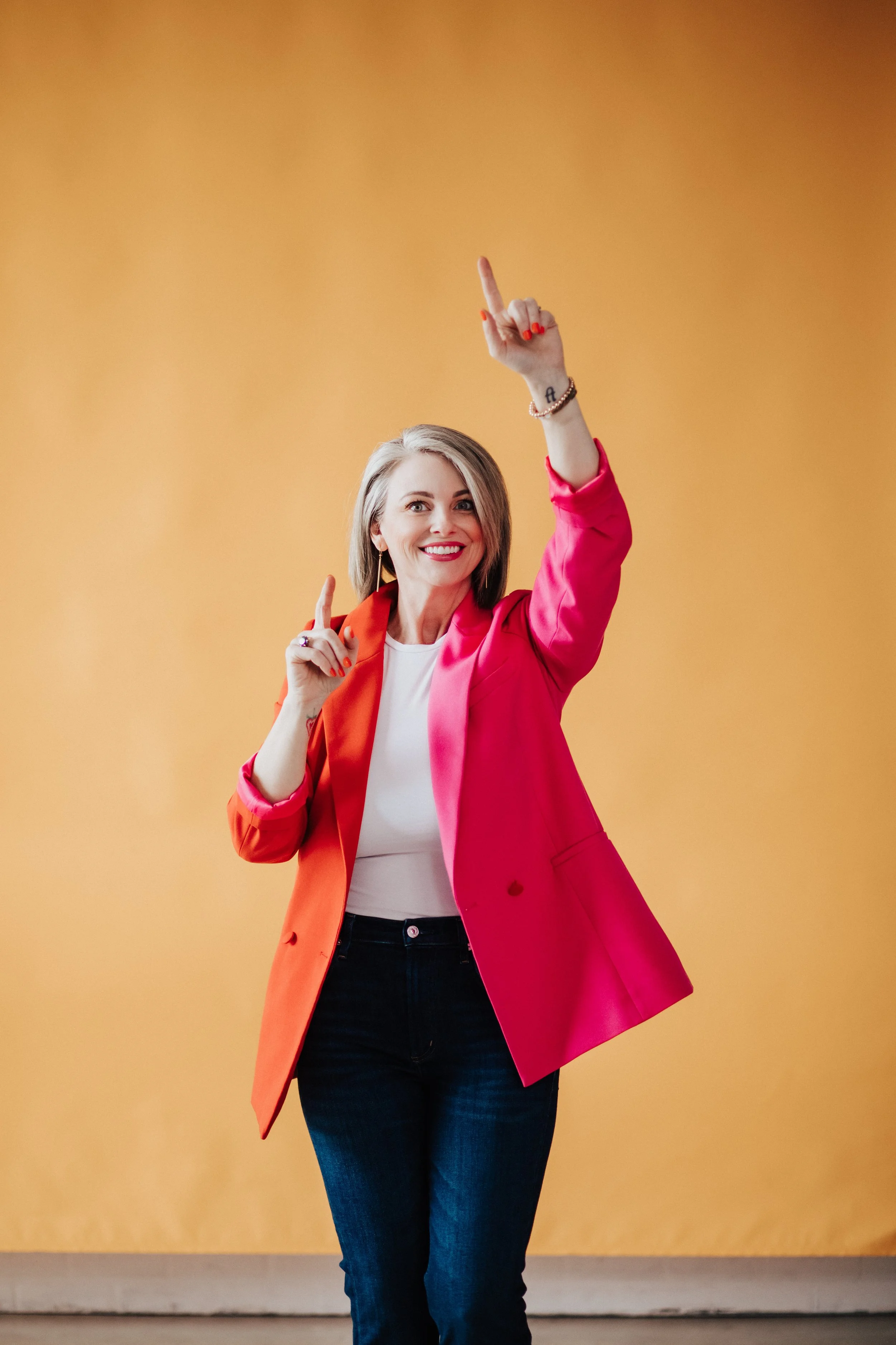 A woman with blonde hair smiling and wearing a pink blazer and dark jeans, holding her right hand with her index finger pointing upwards in front of a plain yellow wall.