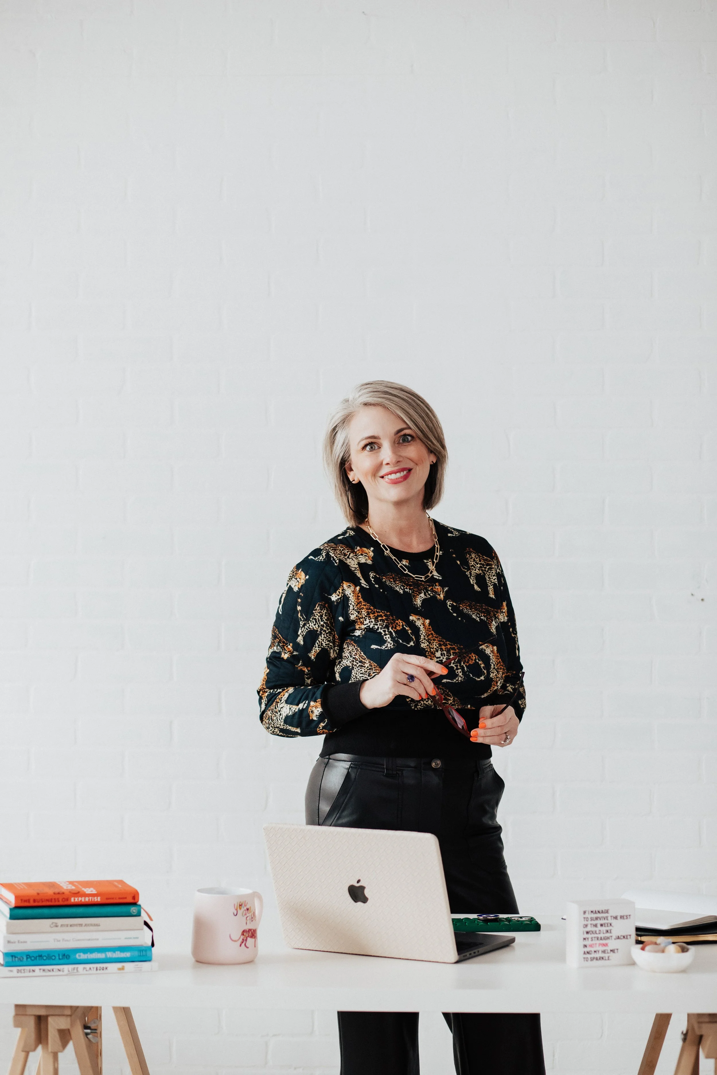 A woman with short blonde hair smiling and holding a pair of glasses stands in a modern office with a white brick wall. There is a white desk in front of her with books, a white mug, a laptop, and other office supplies.