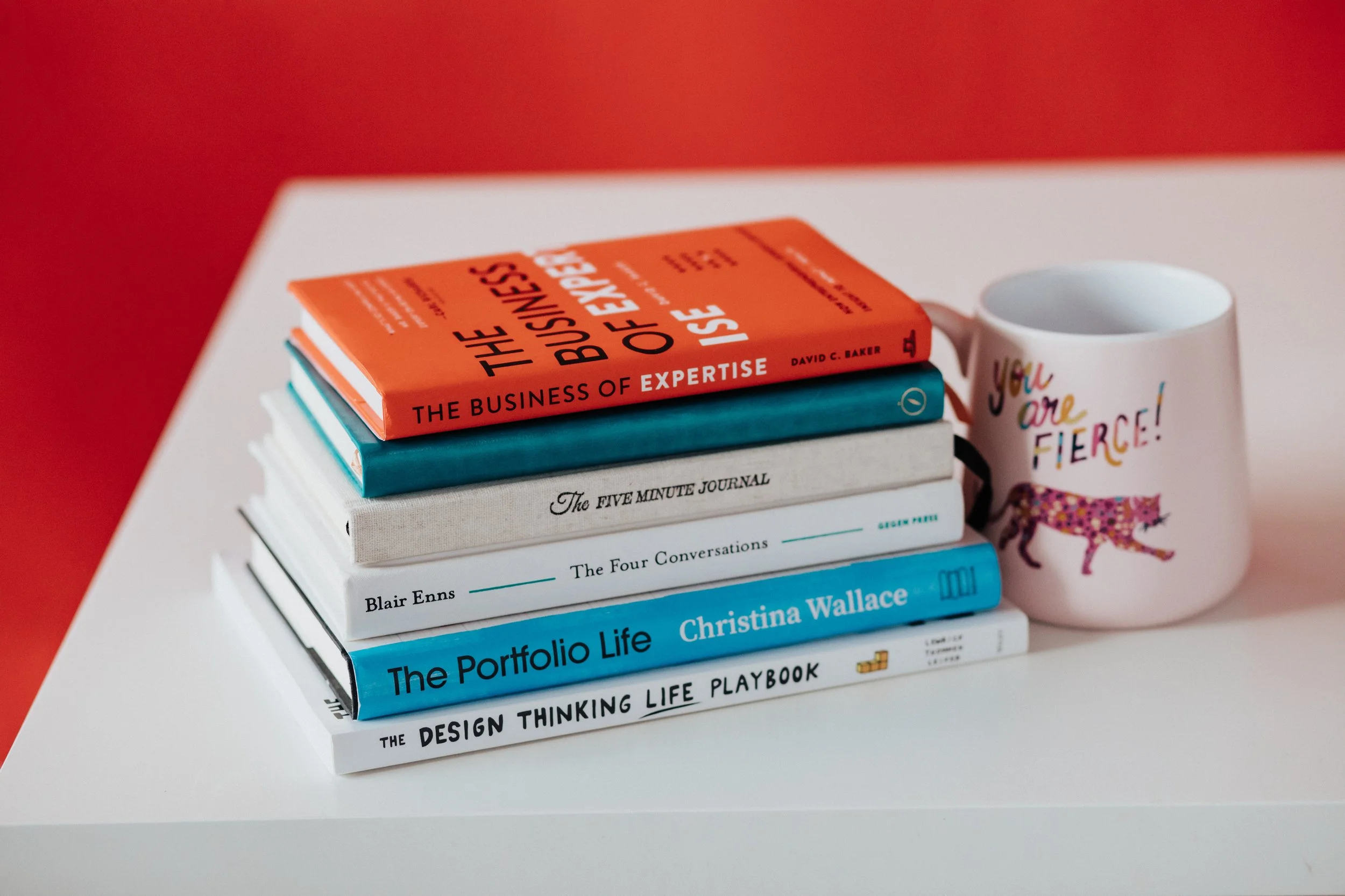 Stack of books and a mug on a white surface. The top book is red and titled 'The Business of Expertise,' next is a white book titled 'The Five Minute Journal,' followed by a blue book titled 'The Four Conversations,' then a teal book titled 'Your Portfolio Life,' and a couple of other books underneath.