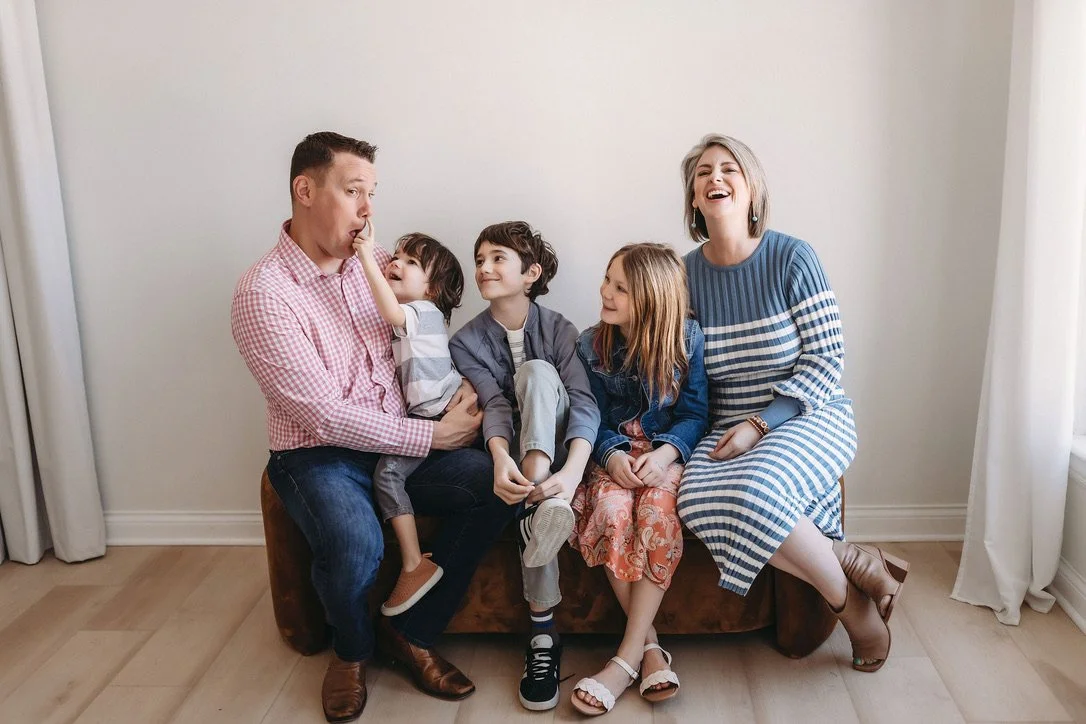 Family of five sitting on a brown couch in a room with light-colored walls and curtains, making funny faces and laughing.