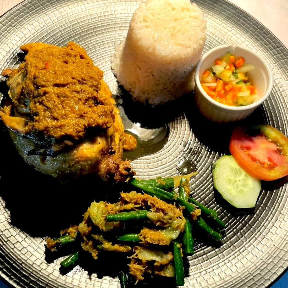A plated meal with rice, a half of chicken betutu topped with yellow sauce, fresh tomato and cucumber slices, a small cup of mixed vegetables, and cooked green bean..
