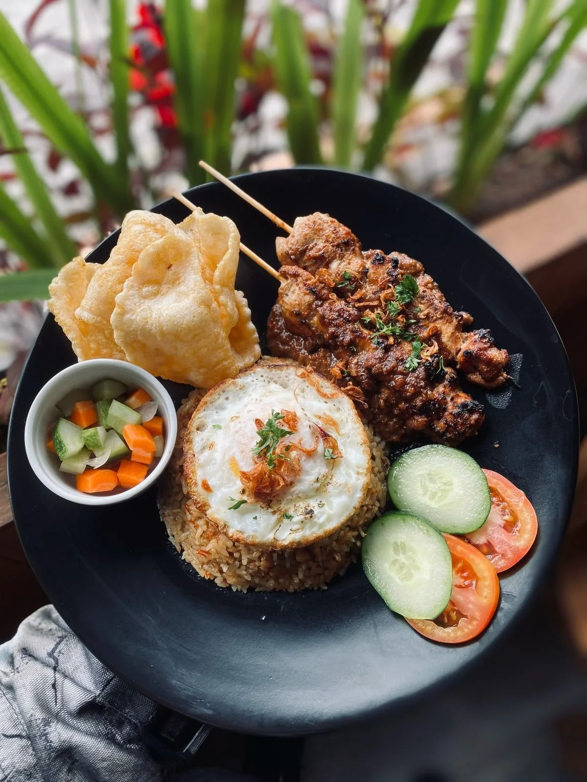Plate of Indonesian food with fried rice, fried egg, satay, cucumber and tomato slices, pickled vegetables, prawn crackers, and a small bowl of pickled cucumbers and carrots.