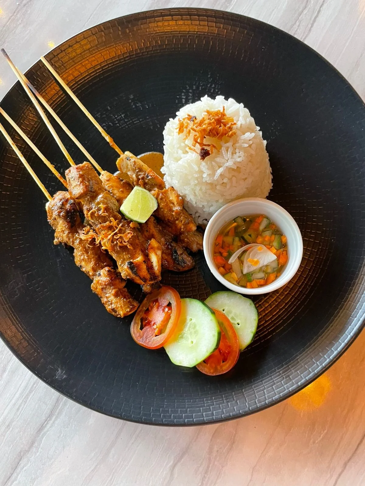 Plate with grilled meat skewers, steamed white rice topped with fried shallots, cucumber and tomato slices, and a small bowl of vegetable soup on a black plate.