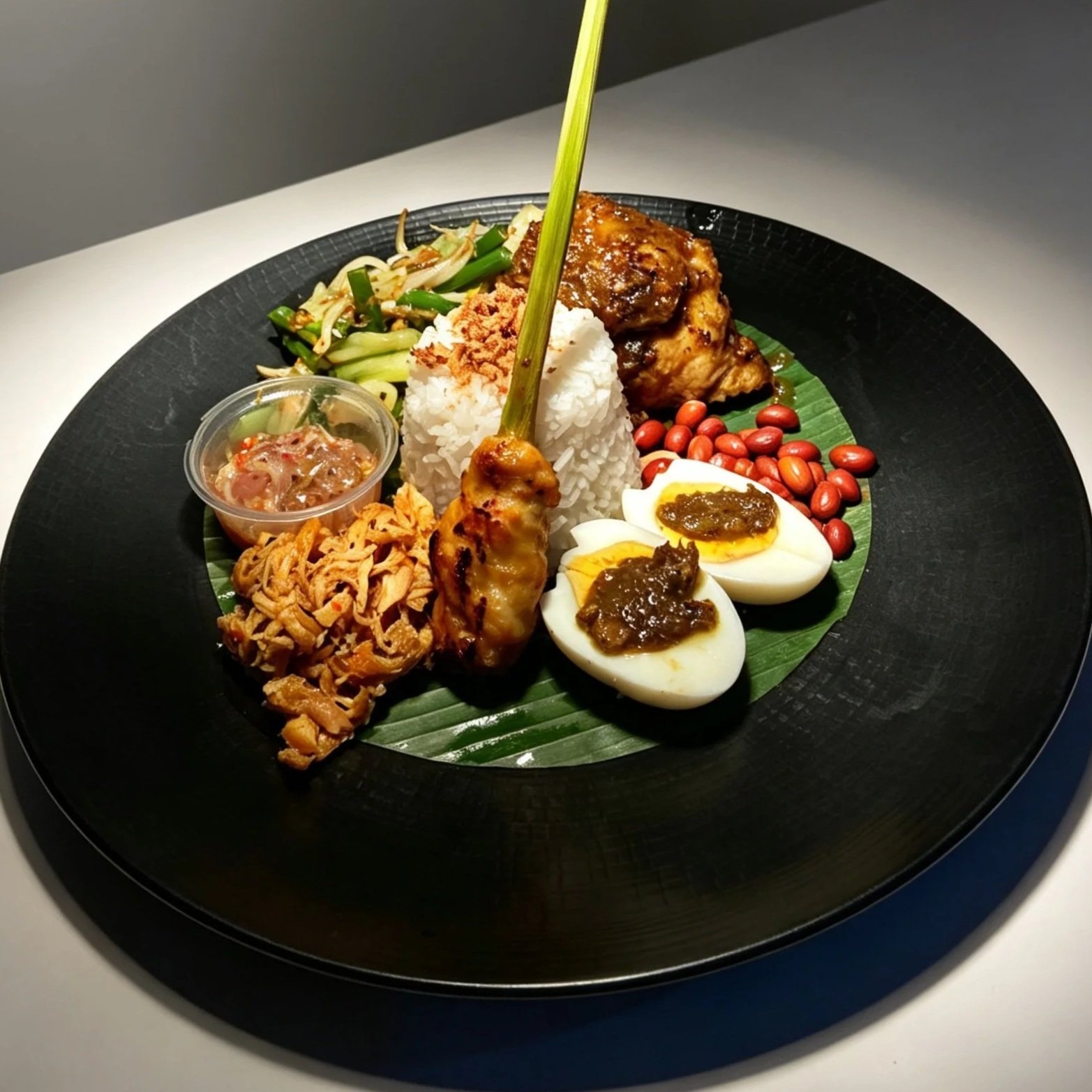 A traditional Balinese plate with rice, sliced boiled eggs topped with sambal, peanuts, fried anchovies, grilled chicken wing, satay skewer, sambal sauce, and vegetable side dishes on a banana leaf.